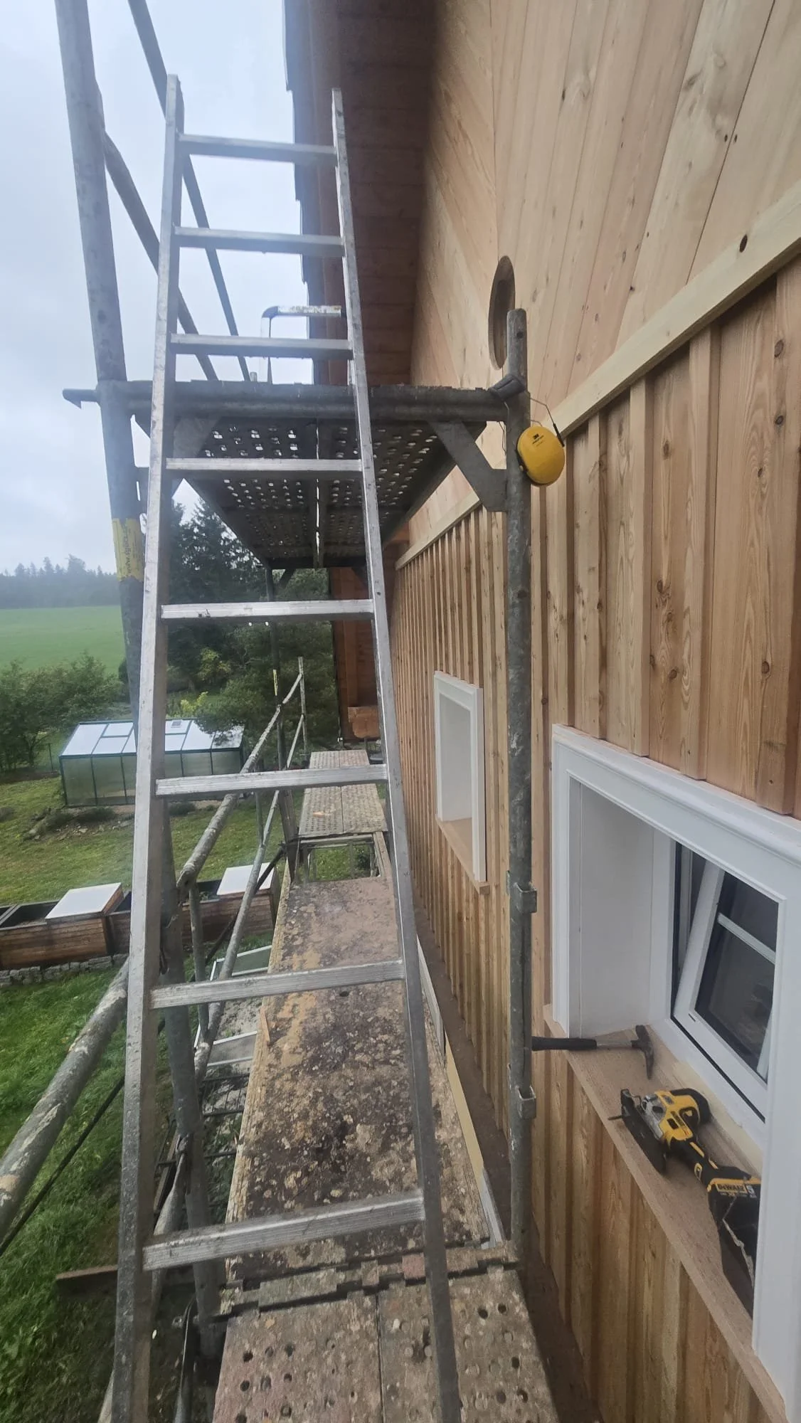 Construction scaffolding on the side of a wooden house with window, tools, and a ladder, overlooking a green yard and garden.