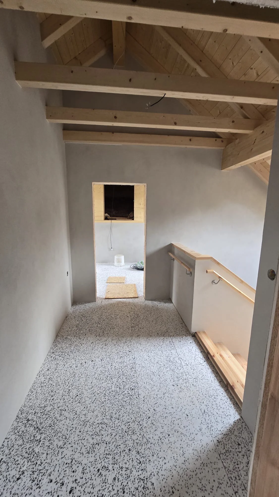 Interior of a house under construction with unfinished walls, wooden ceiling beams, and a staircase railing. The floor has a speckled pattern, and there is an open doorway leading to a small room with construction materials.