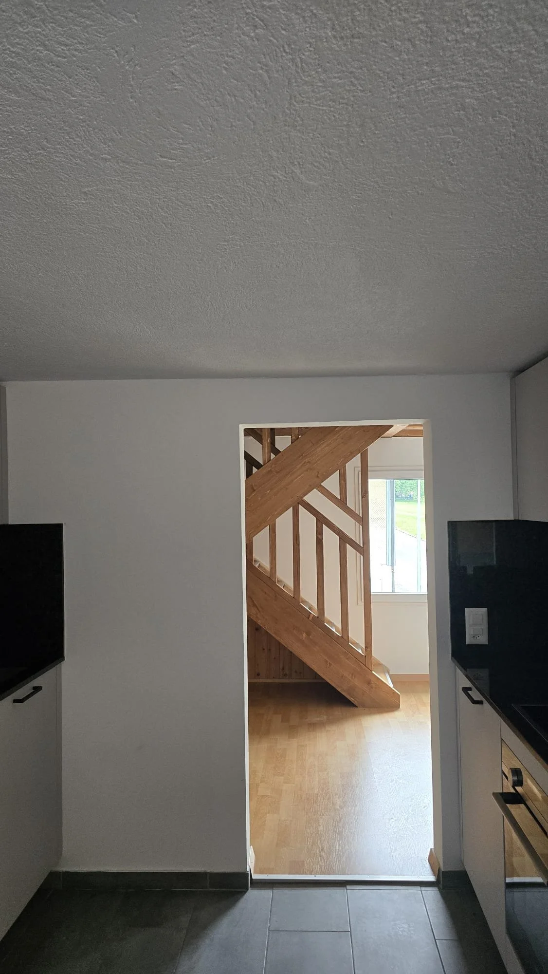 Interior view of a room showing a doorway leading to a space with a wooden staircase, a window, and a wooden floor.