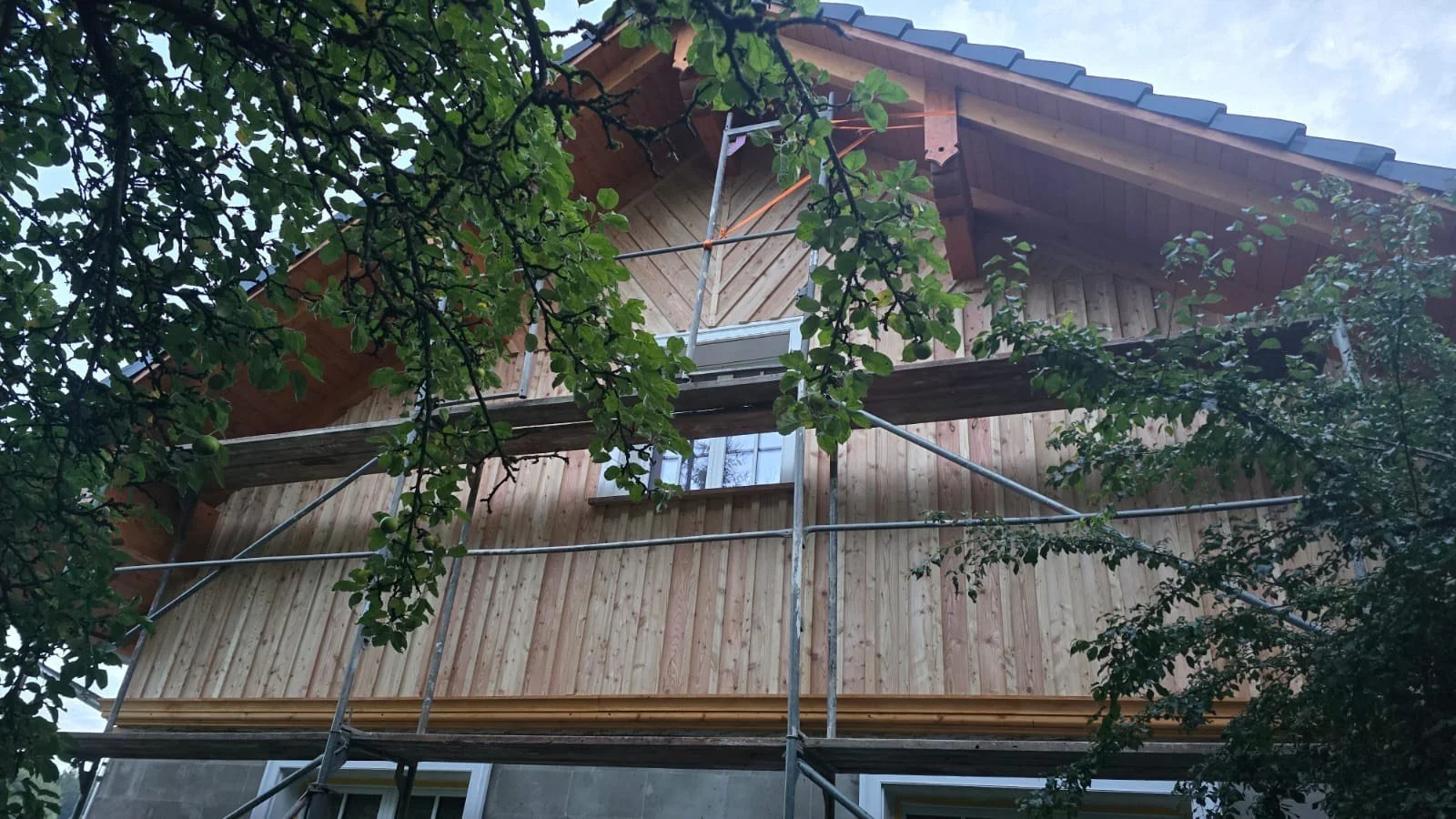 A wooden house under construction with scaffolding around it, surrounded by green trees.