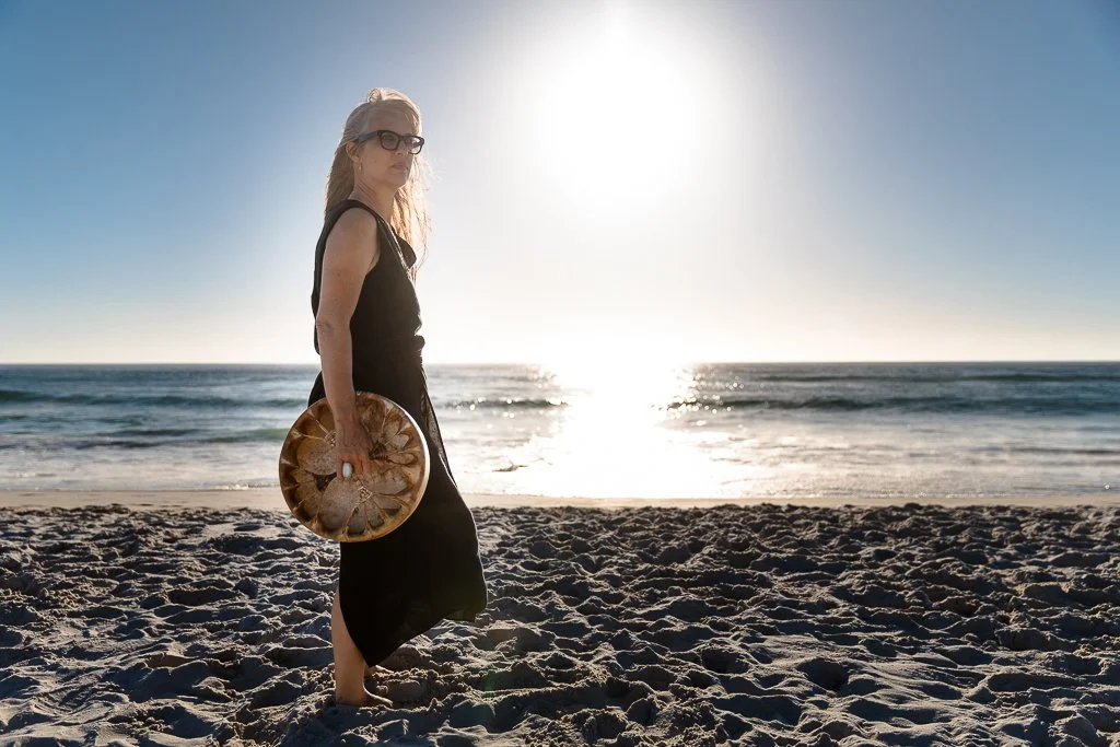 Alrina Laker standing on a beach holding a shamanic drum, representing the depth and precision of The Transformed Leader journey