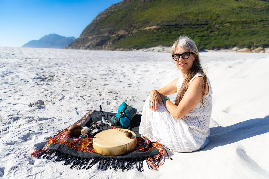 Alrina Laker seated on a beach in Cape Town with her shamanic drum, representing her work with individuals and leaders across the world