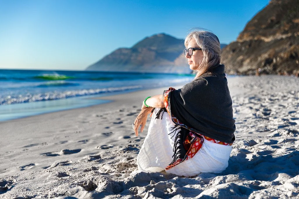 Alrina Laker seated on a beach gazing toward mountains, representing the contemplative inner work at the heart of The Transformed Leader journey