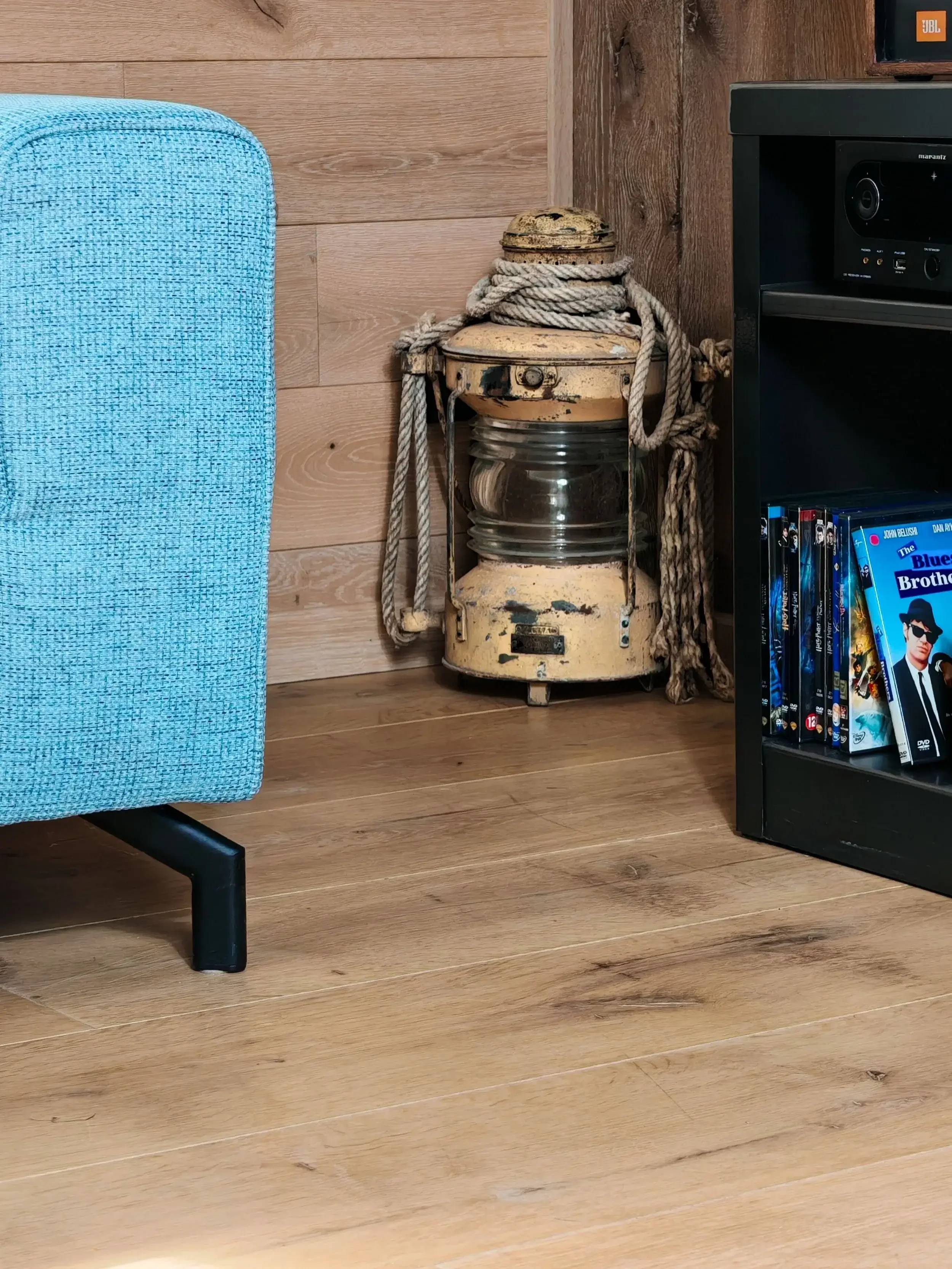 Une vieille lampe de working en métal jaune, entourée de cordes épaisses, posée dans un coin d'une pièce avec un sol en bois et un mur en bois clair.