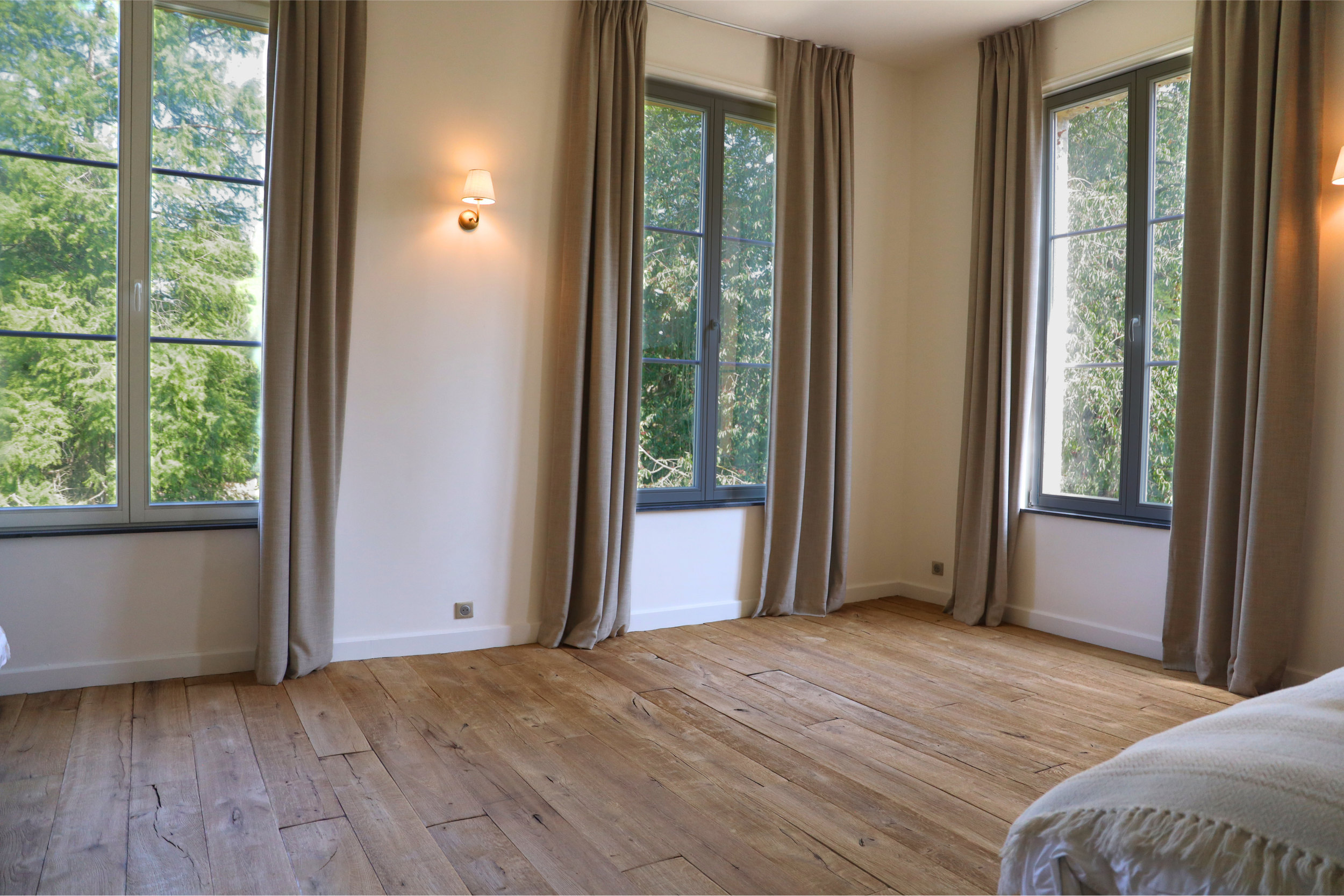 Une chambre chaleureuse avec trois grandes fenêtres laissant entrer beaucoup de lumière naturelle, avec des rideaux beige et un parquet en bois de chêne français texturé et sablé. Typique d'un parquet de Logic Nature Parquets.