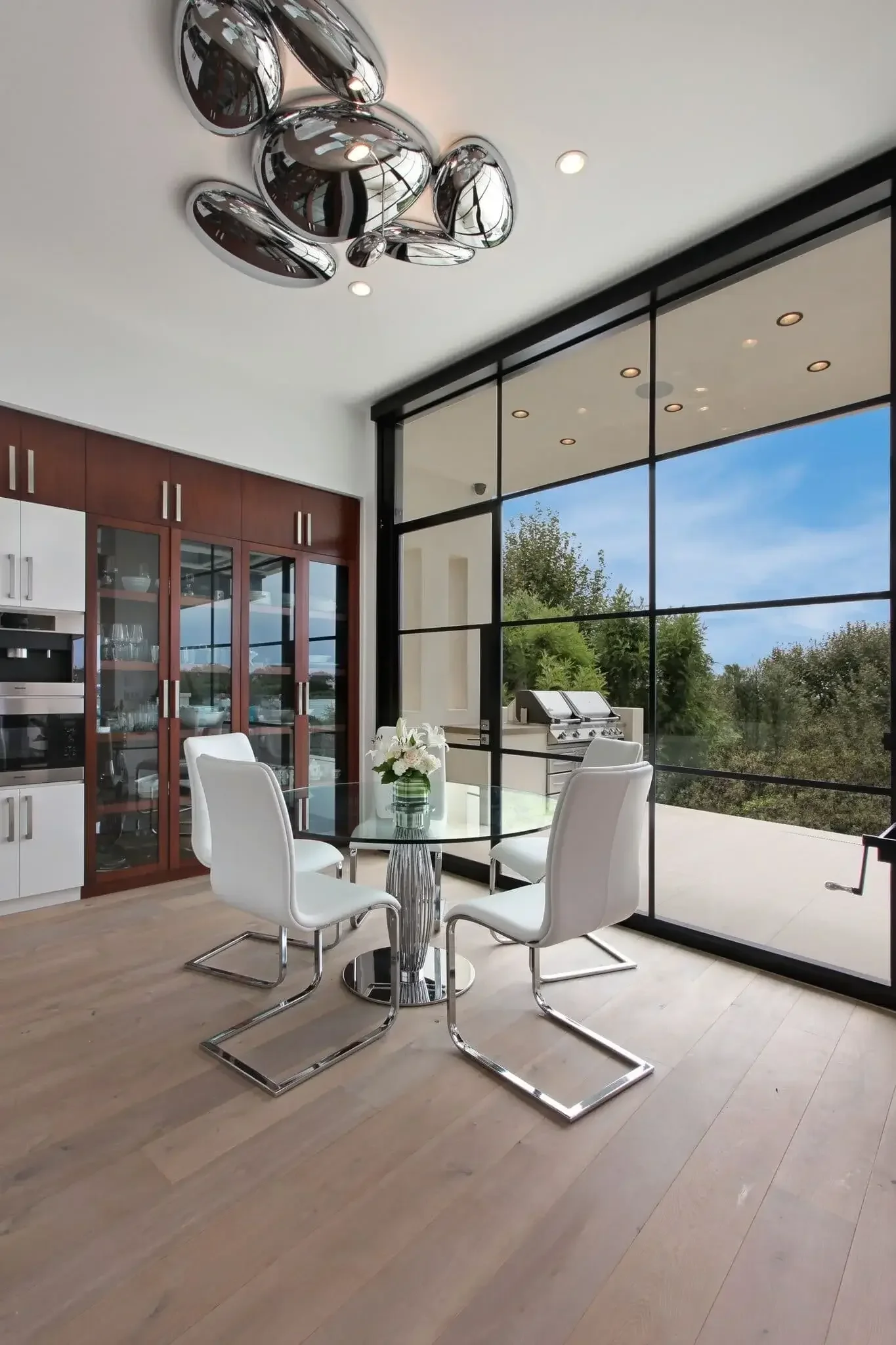 Salle à manger moderne avec une table en verre et quatre chaises blanches, grande baie vitrée avec vue sur un jardin, un barbecue, et un plafonnier métallique.