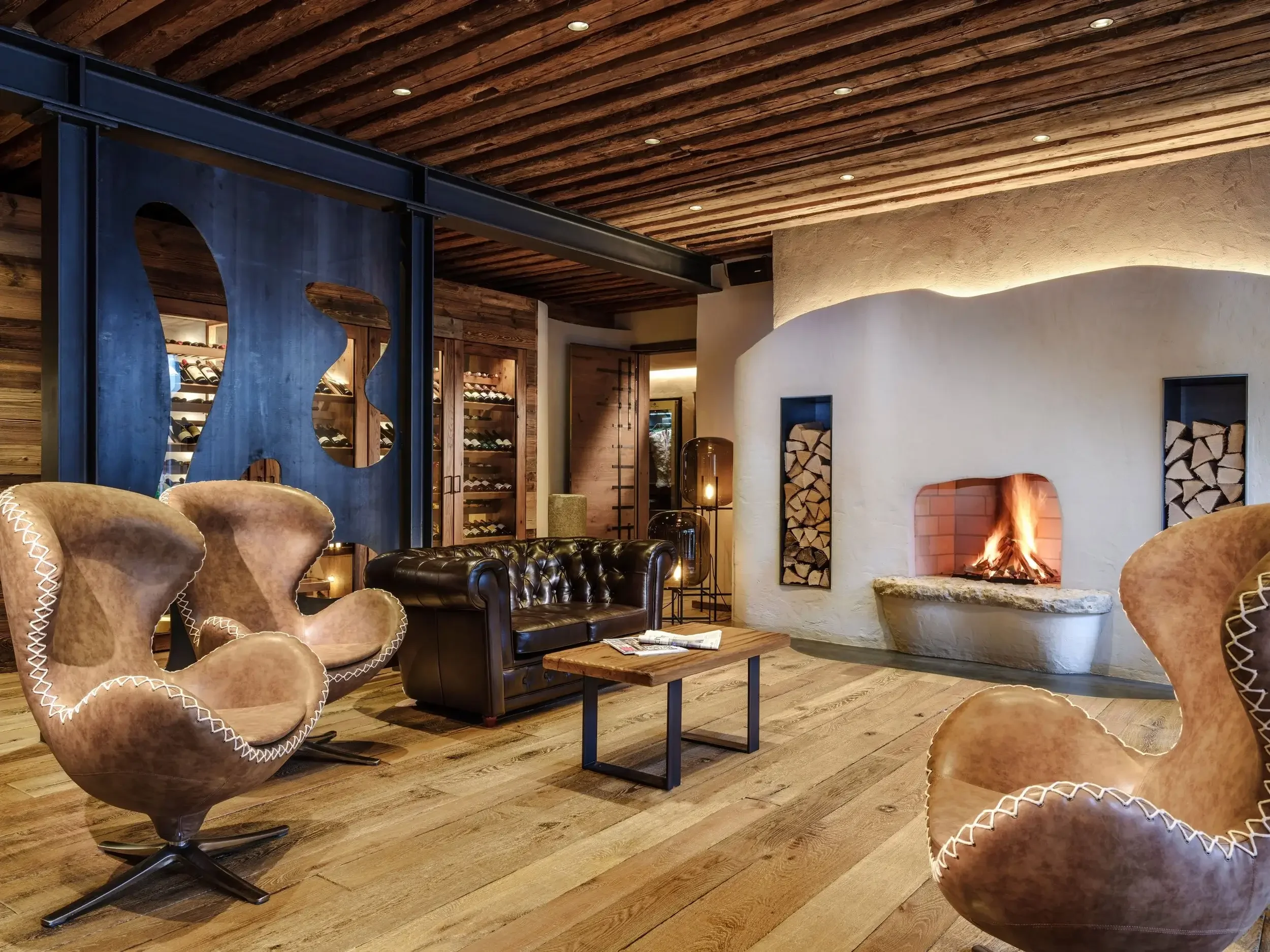 Salon chaleureux avec cheminée, fauteuils en cuir et en velours, étagères en bois avec du bois de chauffage, sol en bois et plafond en bois, décor moderne et cosy.