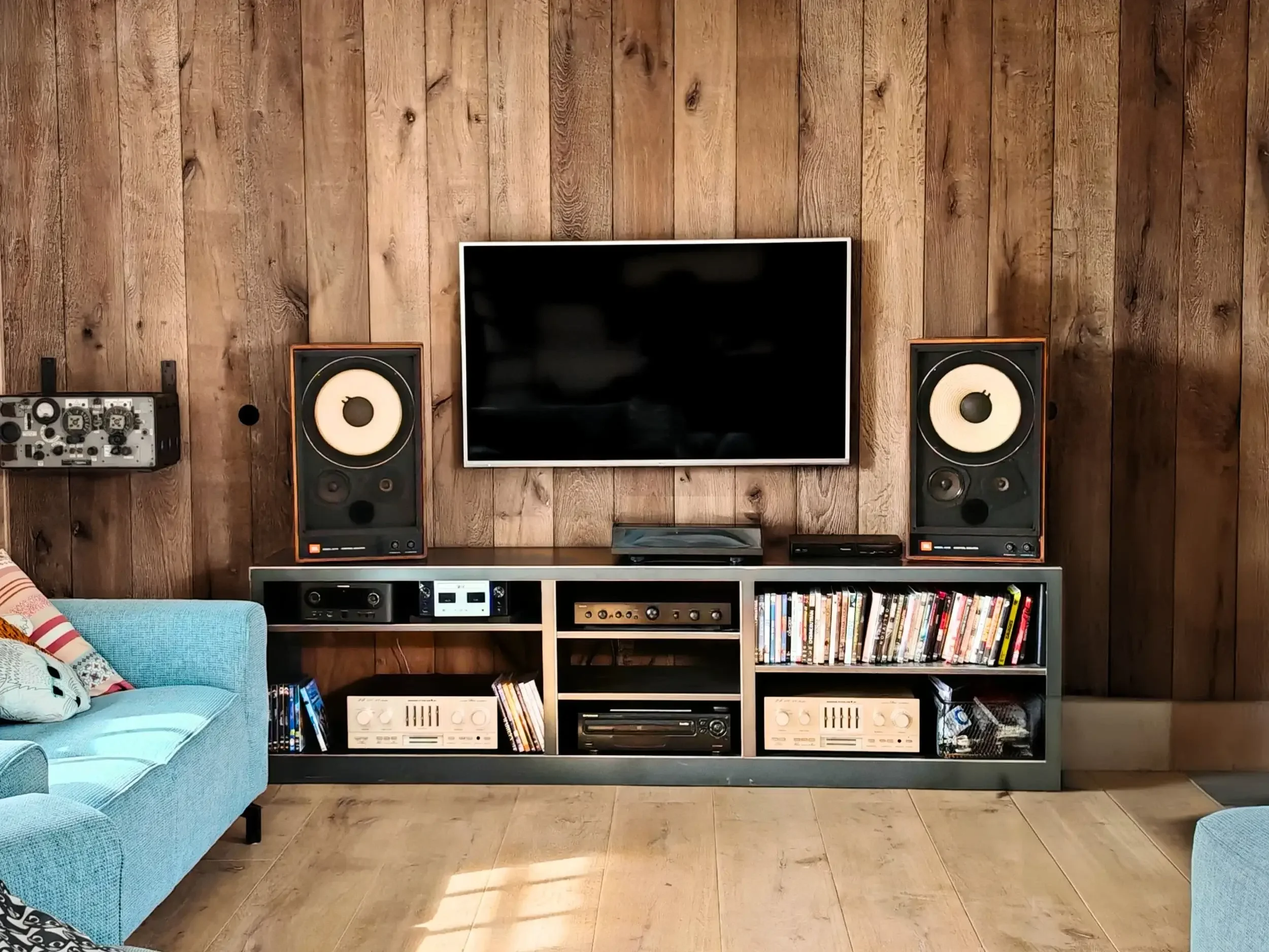 Salon avec un meuble noir contenant une télévision, deux haut-parleurs, une collection de DVDs et de vinyles, un canapé bleu et un mur en chêne massif Weisshorn de la marque Logic Nature Parquets. Au sol un parquet de la gamme Légende clair en chêne.