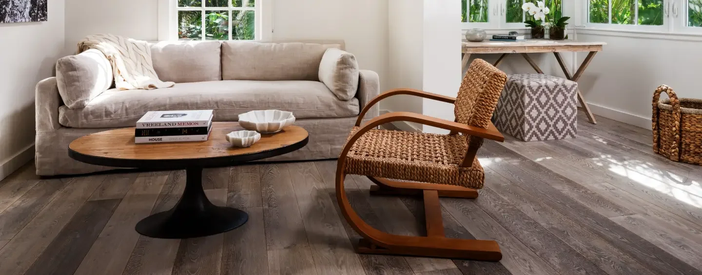 Salon décoratif avec canapé beige, fauteuil en bois et corde, table basse en bois, et vue extérieure à travers les fenêtres.