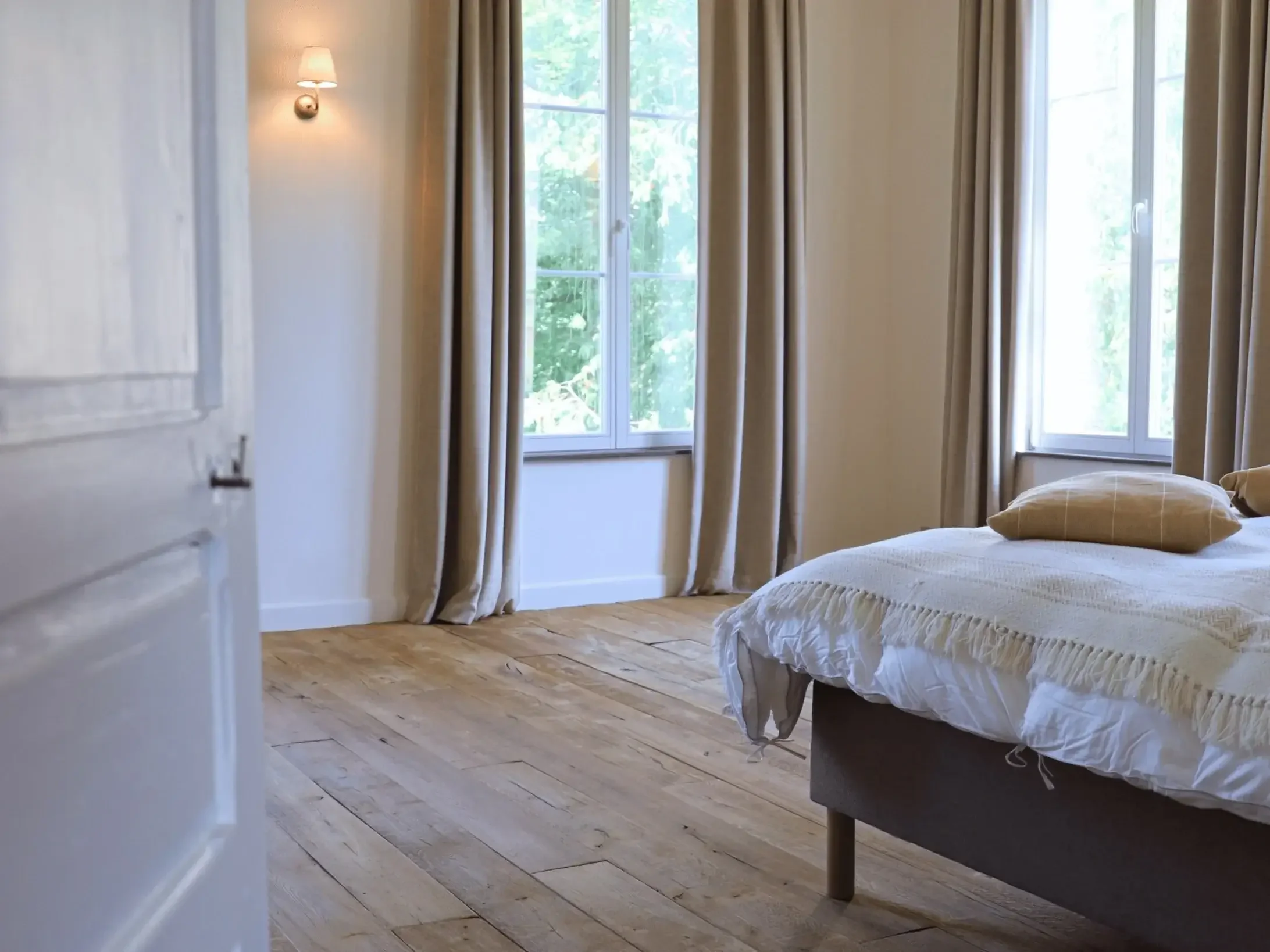 Chambre à coucher lumineuse avec deux fenêtres, un lit fait avec une couverture blanche et des oreillers, rideaux beige et parquet en bois naturel.