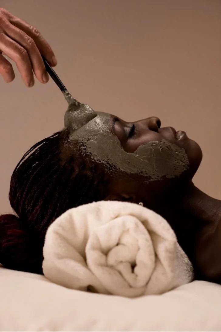 A woman is receiving a facial treatment with mud or clay mask applied to her face, lying down with her head on a rolled white towel.