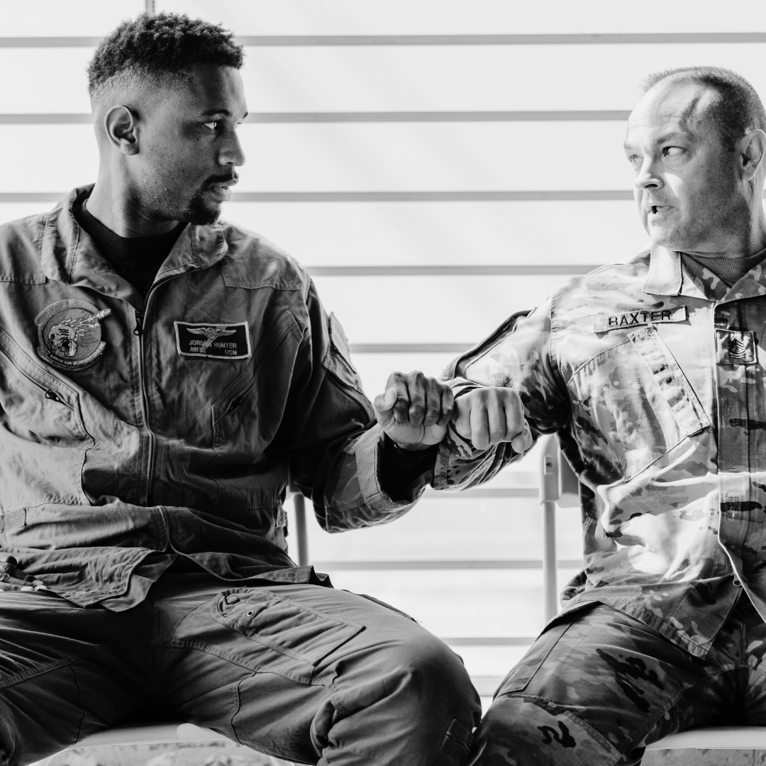 An African American man and a white man, both in military uniform, sitting next to each other and bumping fists.