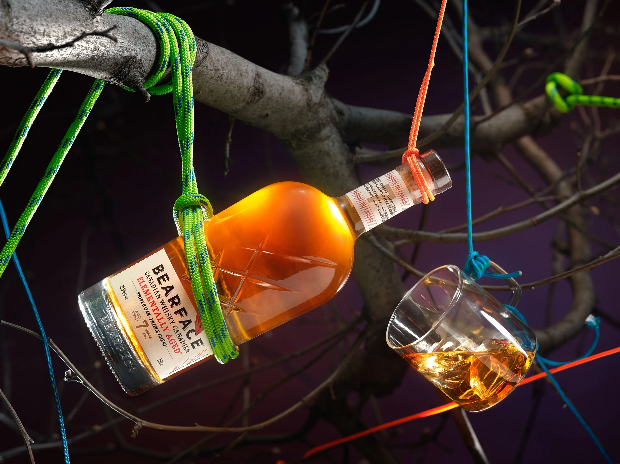 A bottle of Bearface Canadian whisky and a glass of whisky hang from a tree branch, secured with colorful ropes, against a dark background. Chicago Milwaukee Midwest Illinois Food Food beverage Food Drink Savory Photographers photographer photography