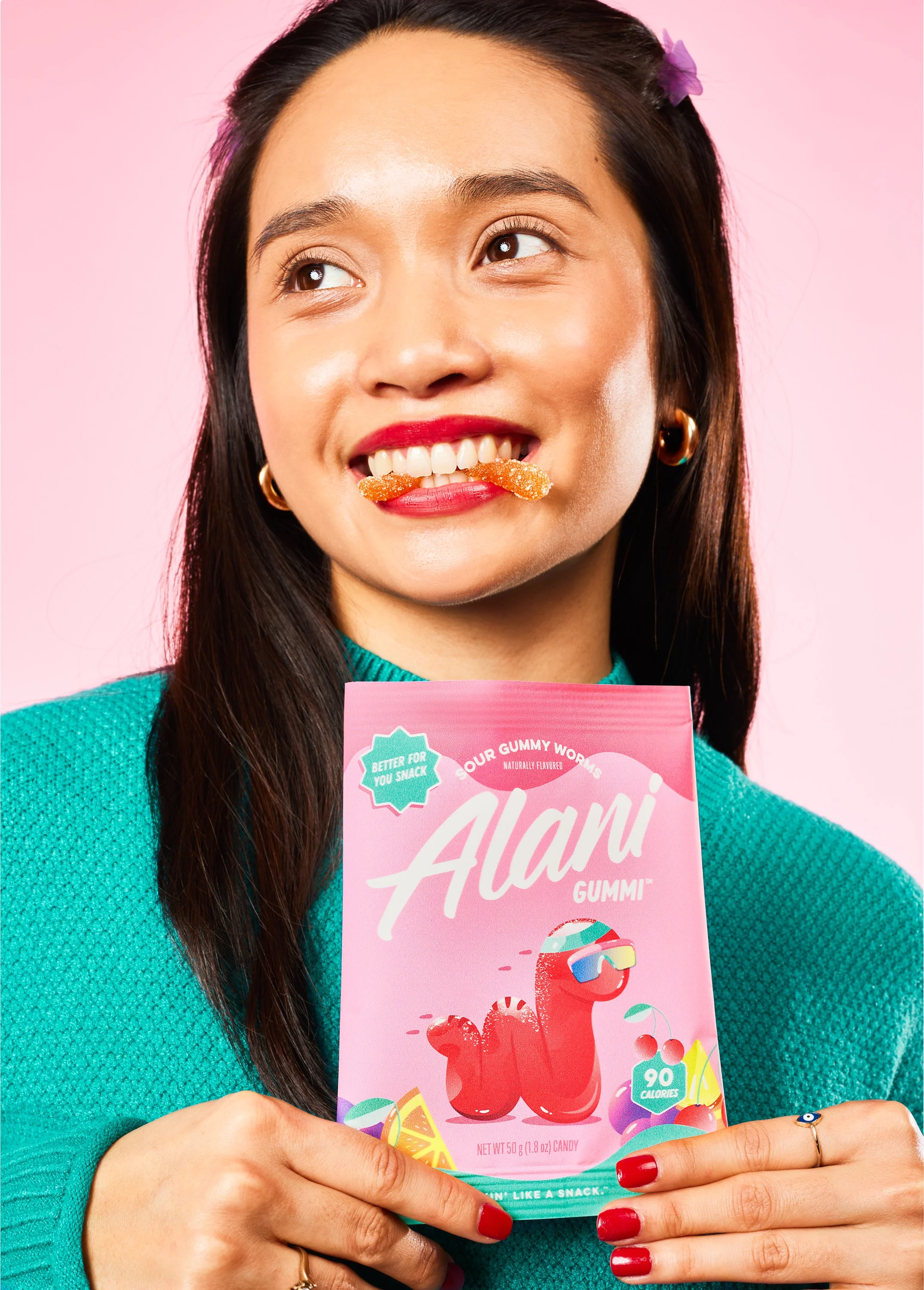 A woman with long dark hair and wearing a teal sweater is holding a pink bag of Alani gummy candies. She has a gummy candy with sugar on her mouth, forming a mustache. She is smiling and looking slightly to the side. The background is pink. Chicago M