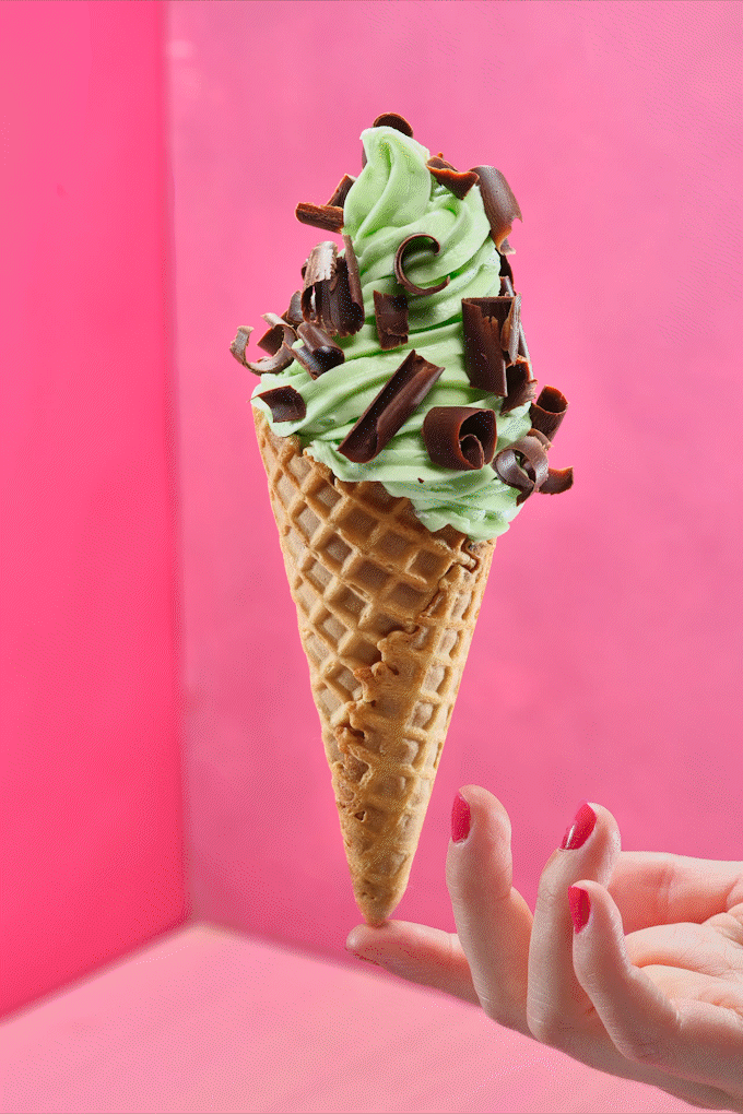 Green soft serve ice cream with chocolate shavings in a waffle cone, held by a hand with pink painted nails against a pink background. Chicago Milwaukee Midwest Illinois Food Food beverage Food Drink Savory Photographers photographer photography vide