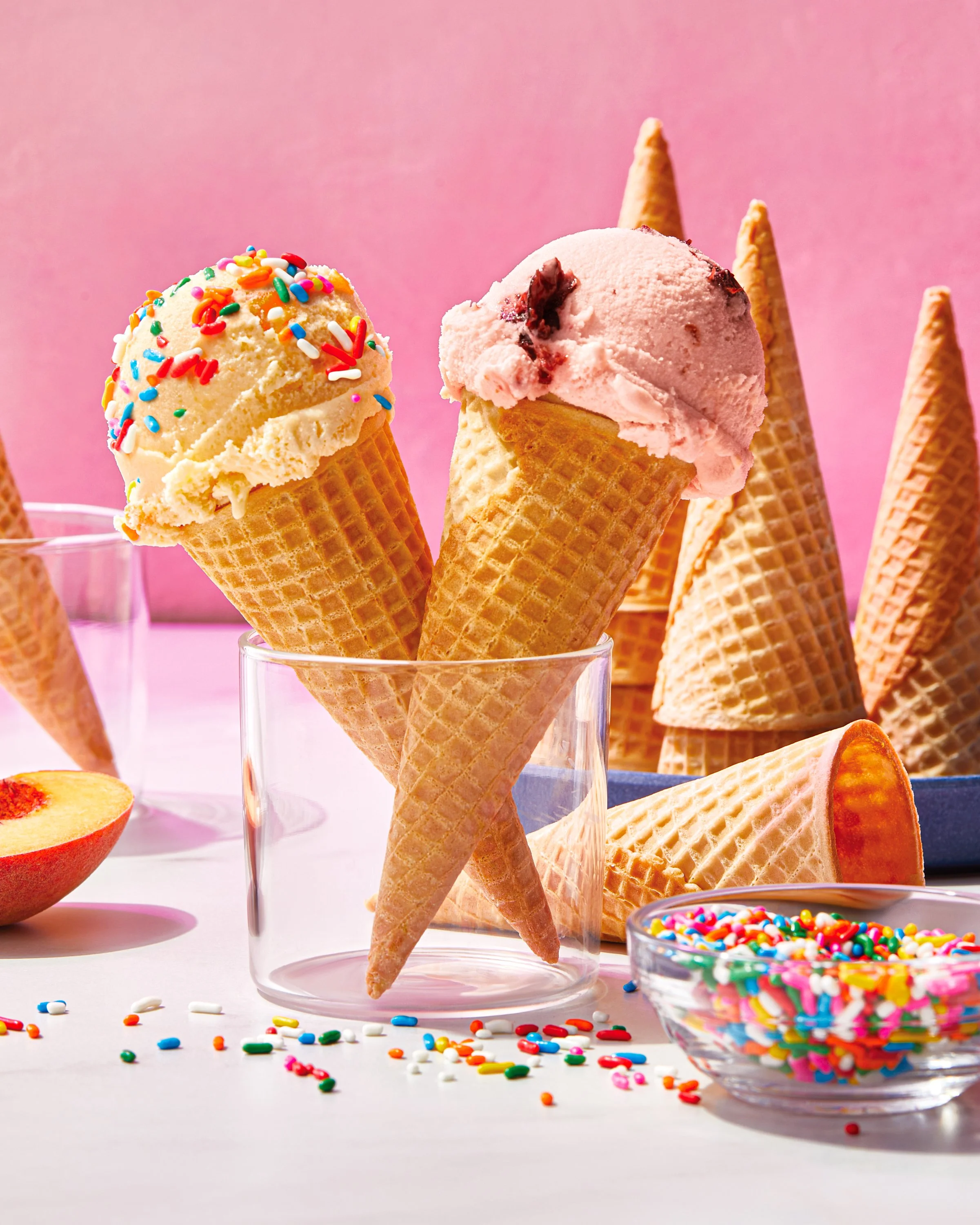Ice cream cones with colorful sprinkles and pink ice cream, decorated with chocolate chunks, surrounded by additional cones, a sliced peach, and a bowl of sprinkles on a pink background. Chicago Milwaukee Midwest Illinois Food Food beverage Food Drin