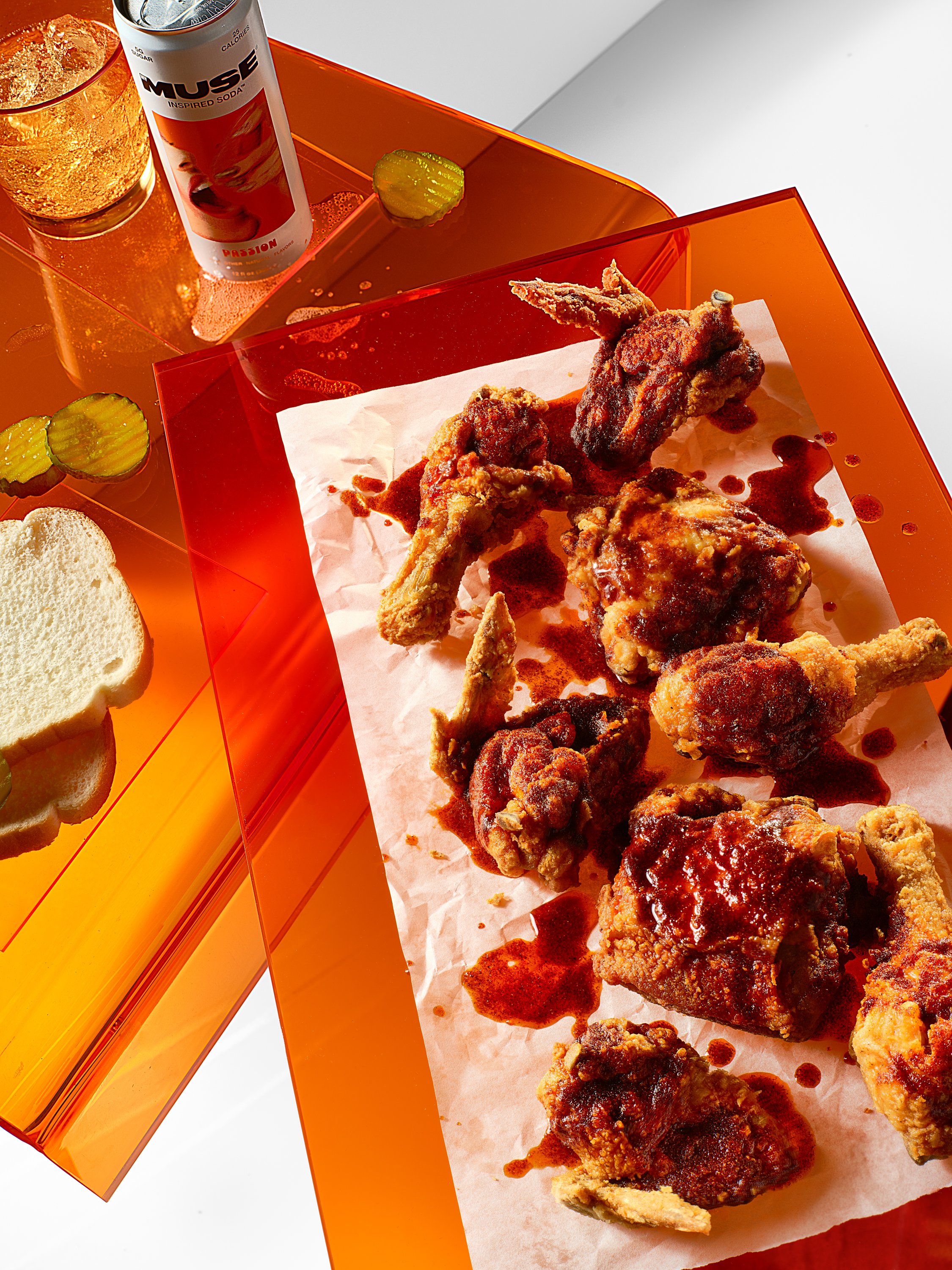 A table featuring fried chicken drumsticks and pieces with barbecue sauce, sliced white bread, pickle slices, a can of Muse Passion soda, and a glass of soda on an orange acrylic table. Chicago Milwaukee Midwest Illinois Food Food beverage Food Drink