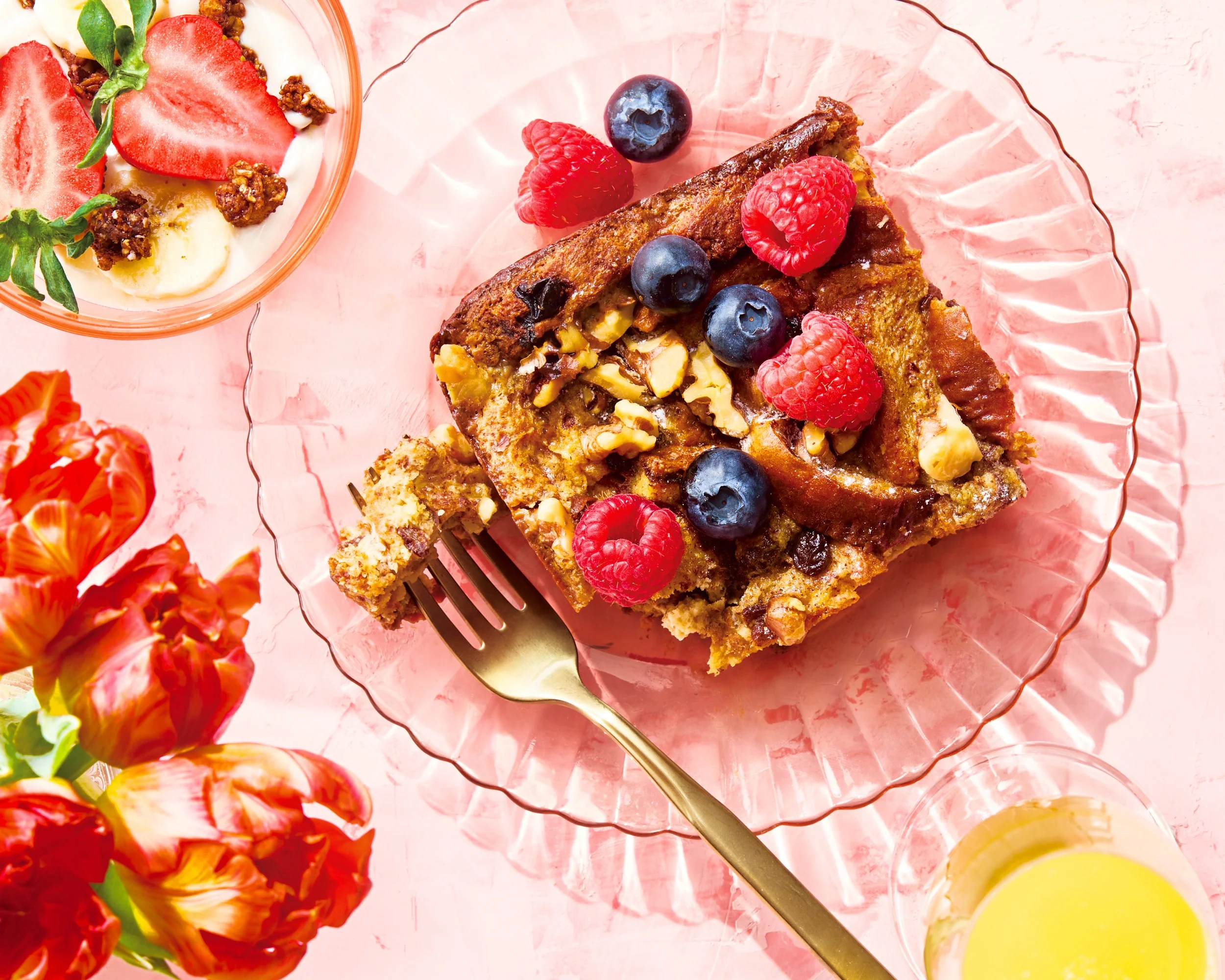 A slice of berry french toast casserole topped with raspberries, blueberries, and chopped nuts on a pink glass plate, with a fork, a bowl of yogurt with strawberries and granola, a glass of lemonade, and orange flowers in the foreground. Chicago Milw