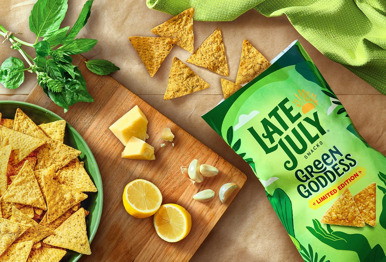 Green bag of Late July Snacks Green Goddess tortilla chips on a table with a green bowl of triangular chips, lemon halves, garlic cloves, cubes of cheese, basil, and a green cloth. Chicago Milwaukee Midwest Illinois Food Food beverage Food Drink Savo