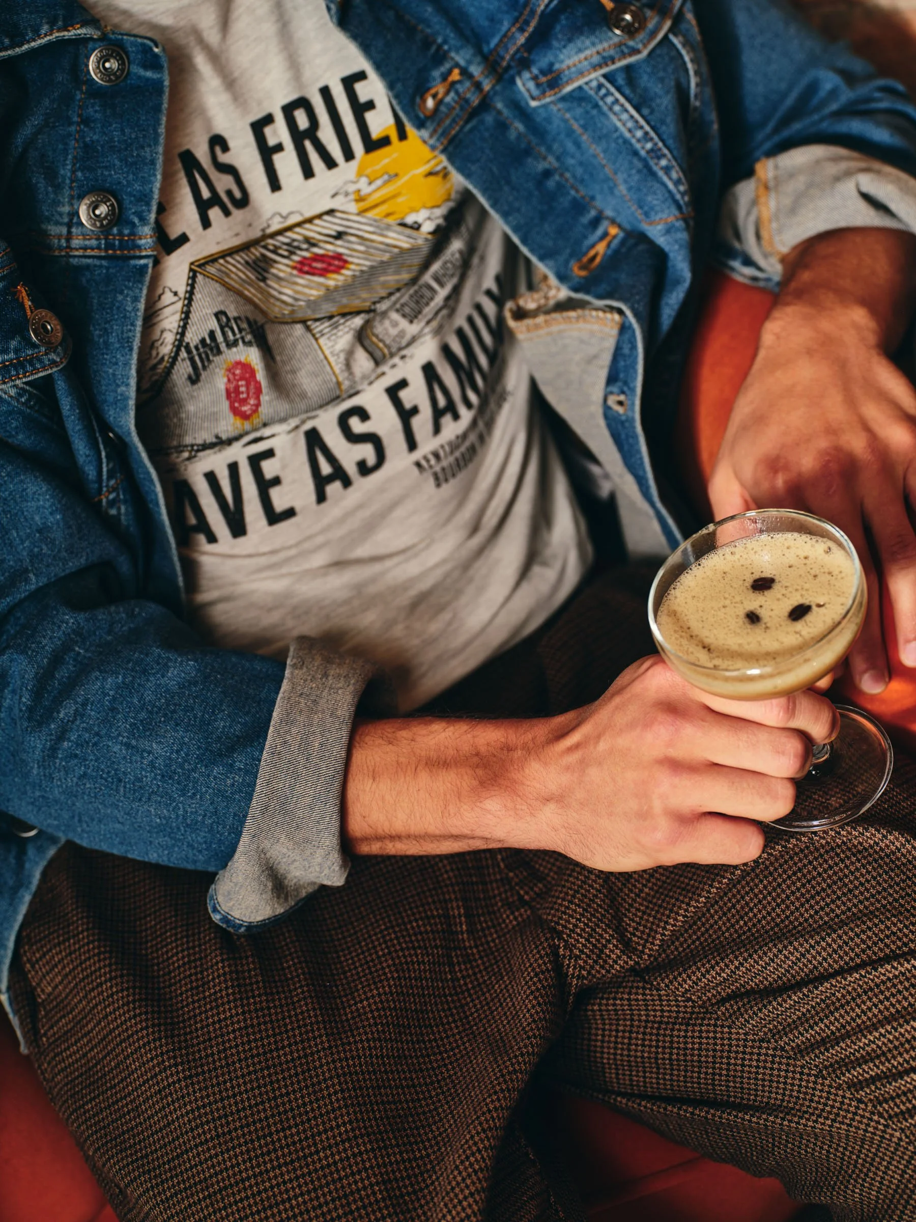 Person wearing a denim jacket and graphic t-shirt, holding a coffee cocktail in a glass cup.