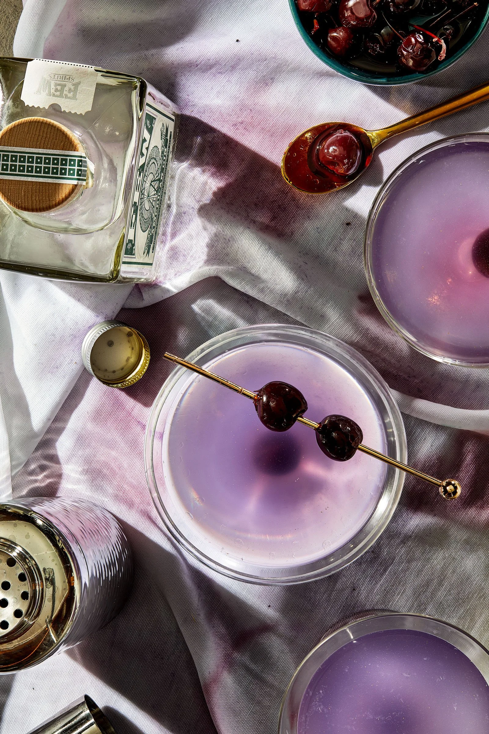 Beverages with purple liquid and cherries, a bowl of dark cherries, a bottle of alcohol with a wafer cookie, a cocktail shaker, and a gold spoon with cherries on top, on a pink tablecloth. Chicago Milwaukee Midwest Illinois Food Food beverage Food Dr