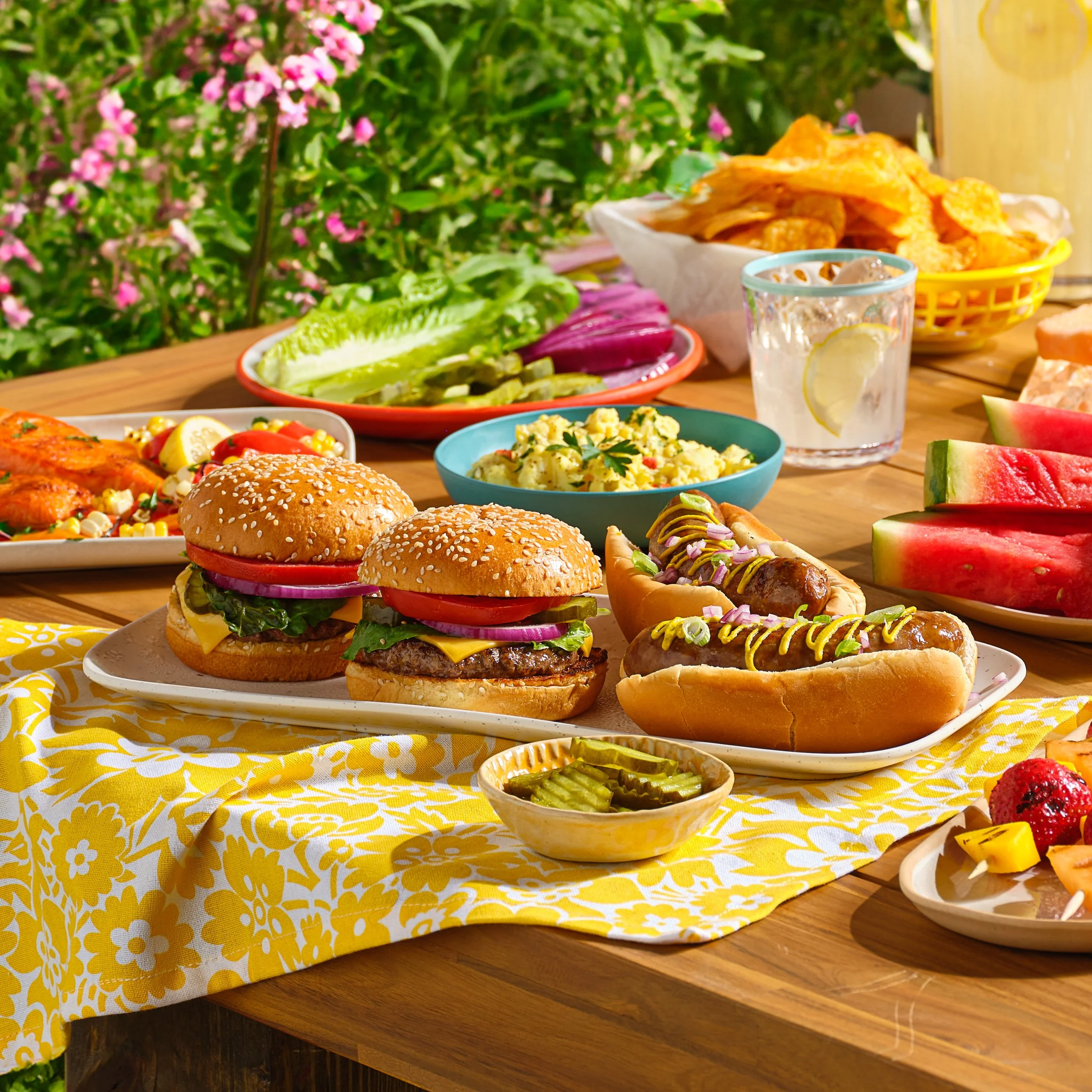 A colorful outdoor picnic table with various foods including mini cheeseburgers, hot dogs, watermelon slices, potato chips, potato salad, pickles, vegetables, and drinks Chicago Milwaukee Midwest Illinois Food Food beverage Food Drink Savory Photogra