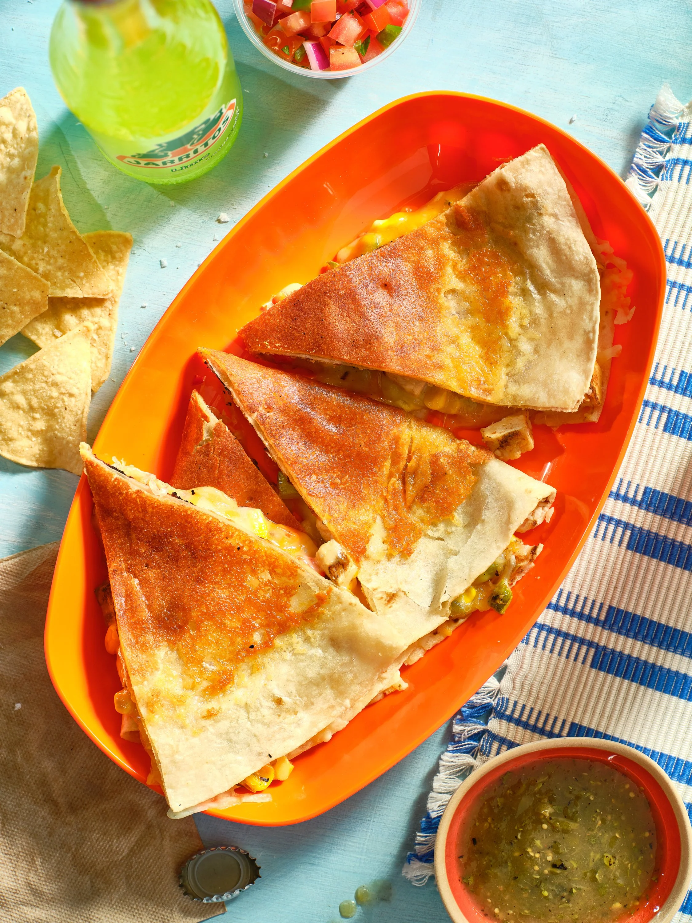 Three slices of quesadilla on an orange plate, accompanied by a small bowl of green salsa, tortilla chips, a green bottle, and a bowl of pico de gallo on a light blue table. Chicago Milwaukee Midwest Illinois Food Food beverage Food Drink Savory Phot