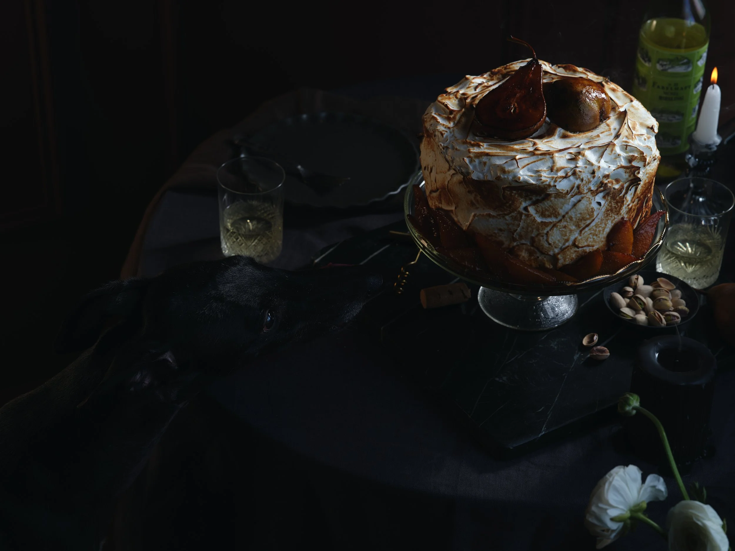 A dark table setting with a large cake topped with pears and a meringue, surrounded by glasses, pistachillos, and flowers, with a black dog licking the table. Chicago Milwaukee Midwest Illinois Food Food beverage Food Drink Savory Photographers photo