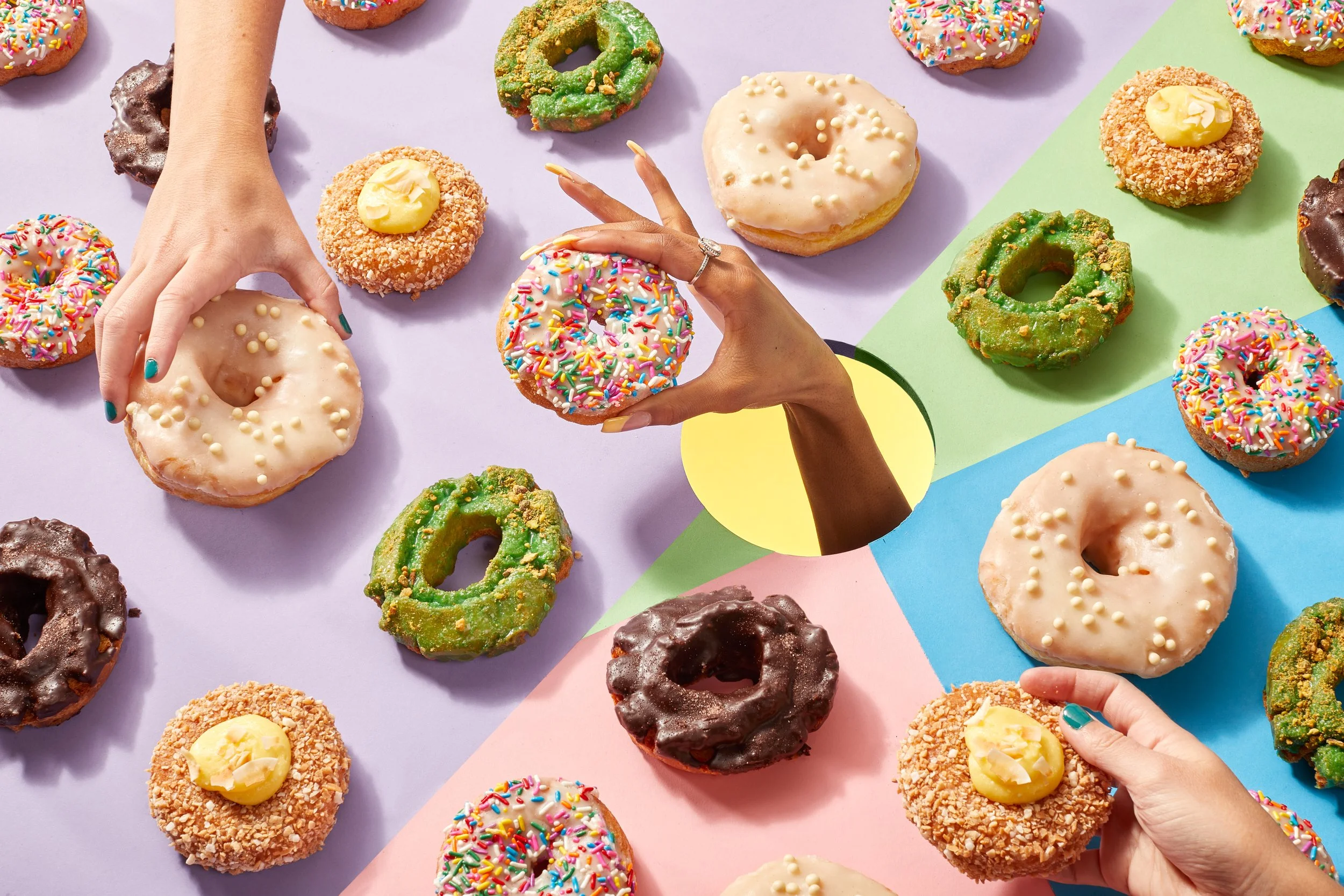 Colorful donuts with sprinkles, icing, and toppings spread on pastel-colored background, with hands reaching to pick them up. Chicago Milwaukee Midwest Illinois Food Food beverage Food Drink Savory Photographers photographer photography videography v