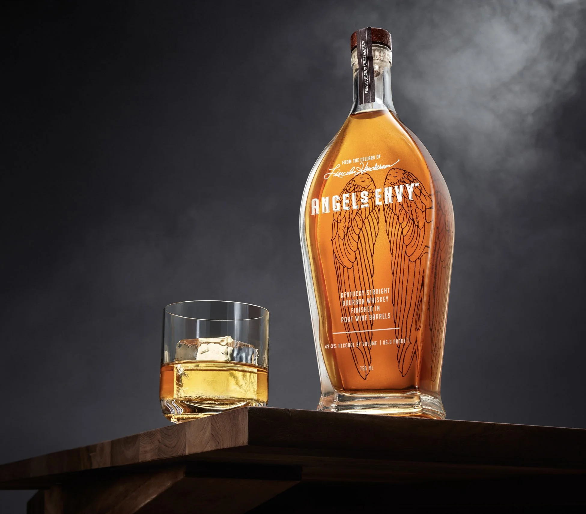 A bottle of Angels Envy Kentucky straight bourbon whiskey with a glass of whiskey with ice next to it, on a wooden surface against a dark gray background. Chicago Milwaukee Midwest Illinois Food Food beverage Food Drink Savory Photographers photograp