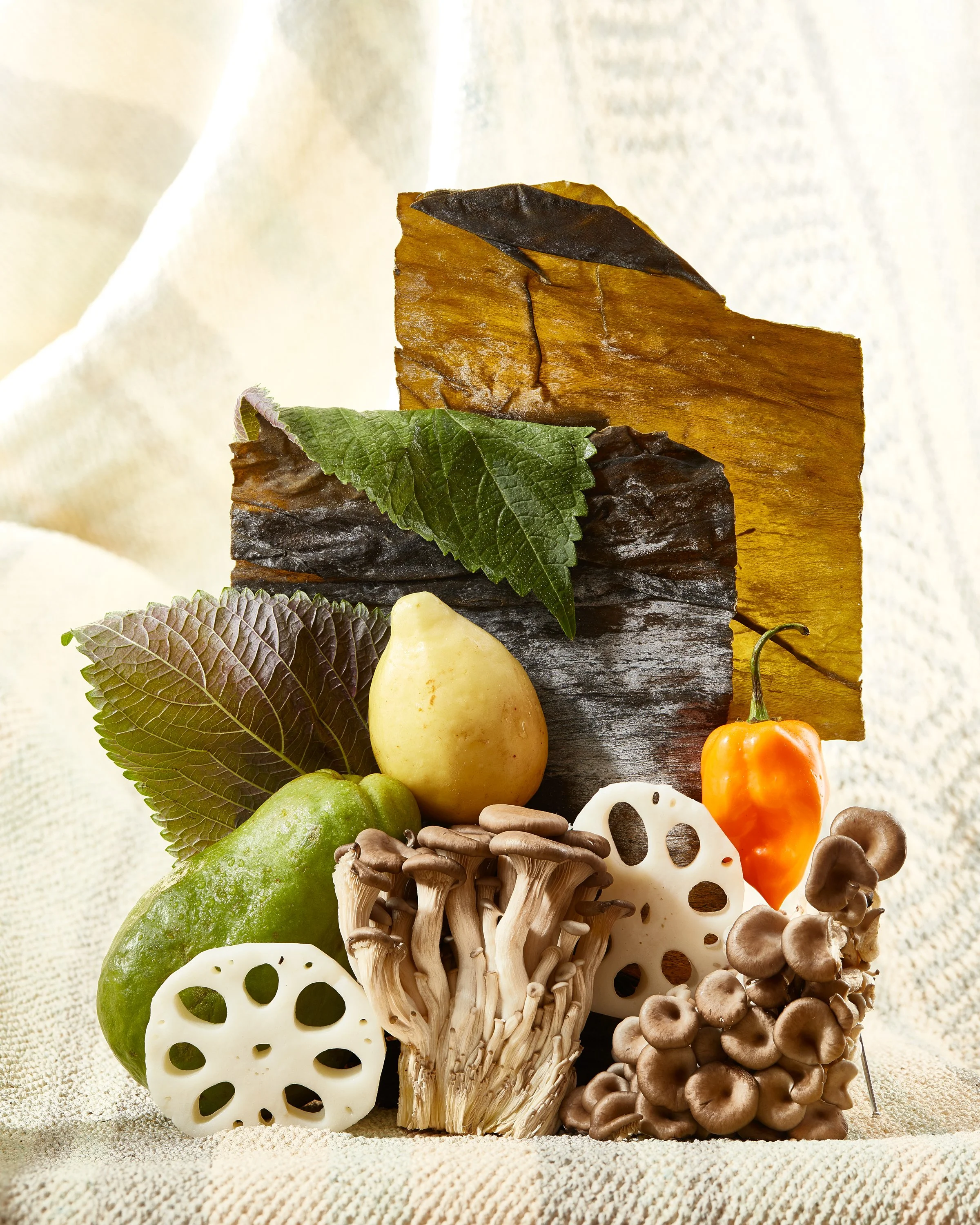 Collection of various vegetables and fungi on a light fabric background, including lotus root, enoki mushrooms, ginkgo nuts, a green pepper, a yellow pear-shaped fruit, an orange pepper, and large green and purple leaves. Chicago Milwaukee Midwest Il