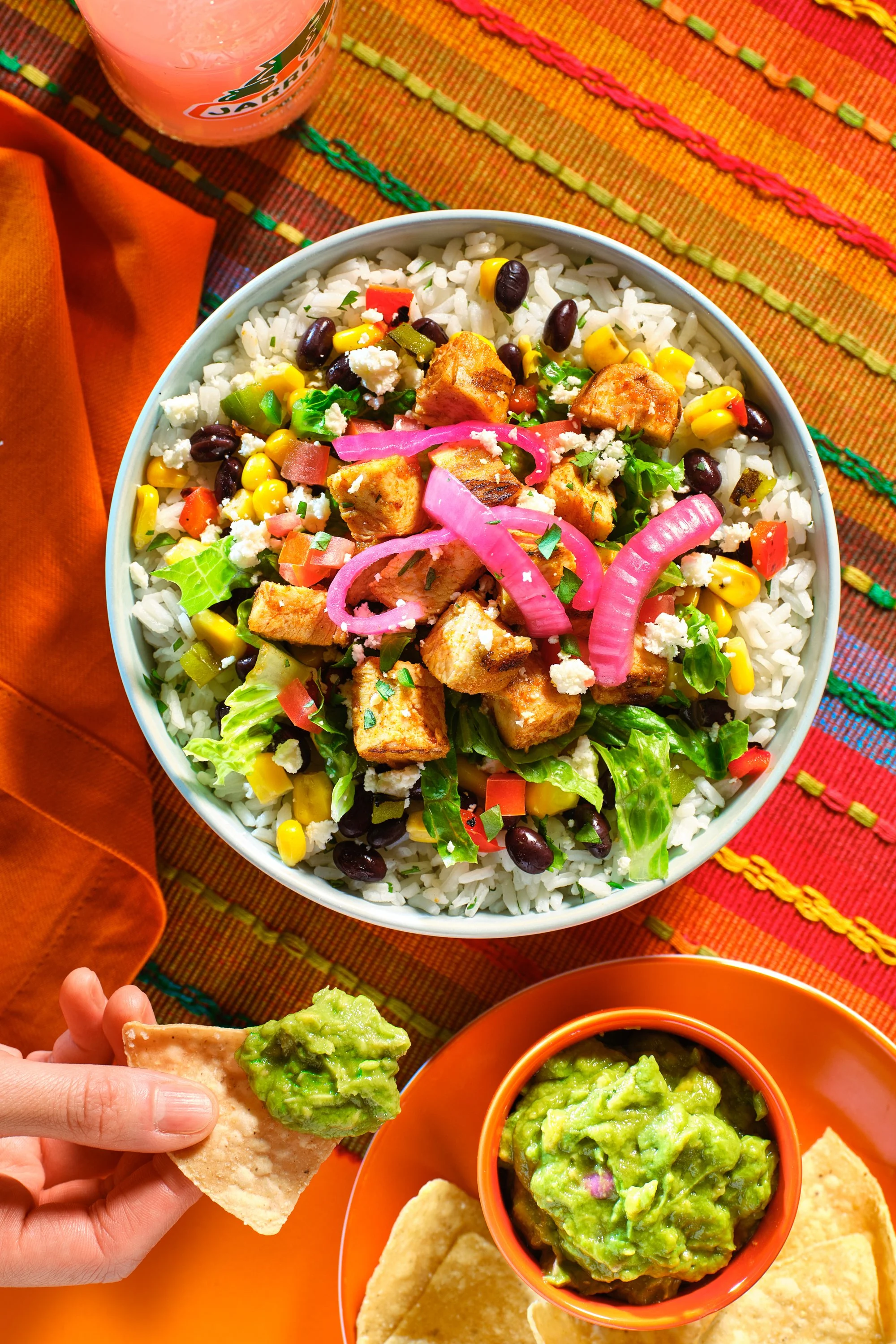 A colorful Mexican-style meal with a bowl of rice topped with grilled chicken, black beans, corn, lettuce, diced tomatoes, pickled onions, and crumbled cheese. Beside it is a plate with guacamole and tortilla chips, and a small bowl of extra guacamol