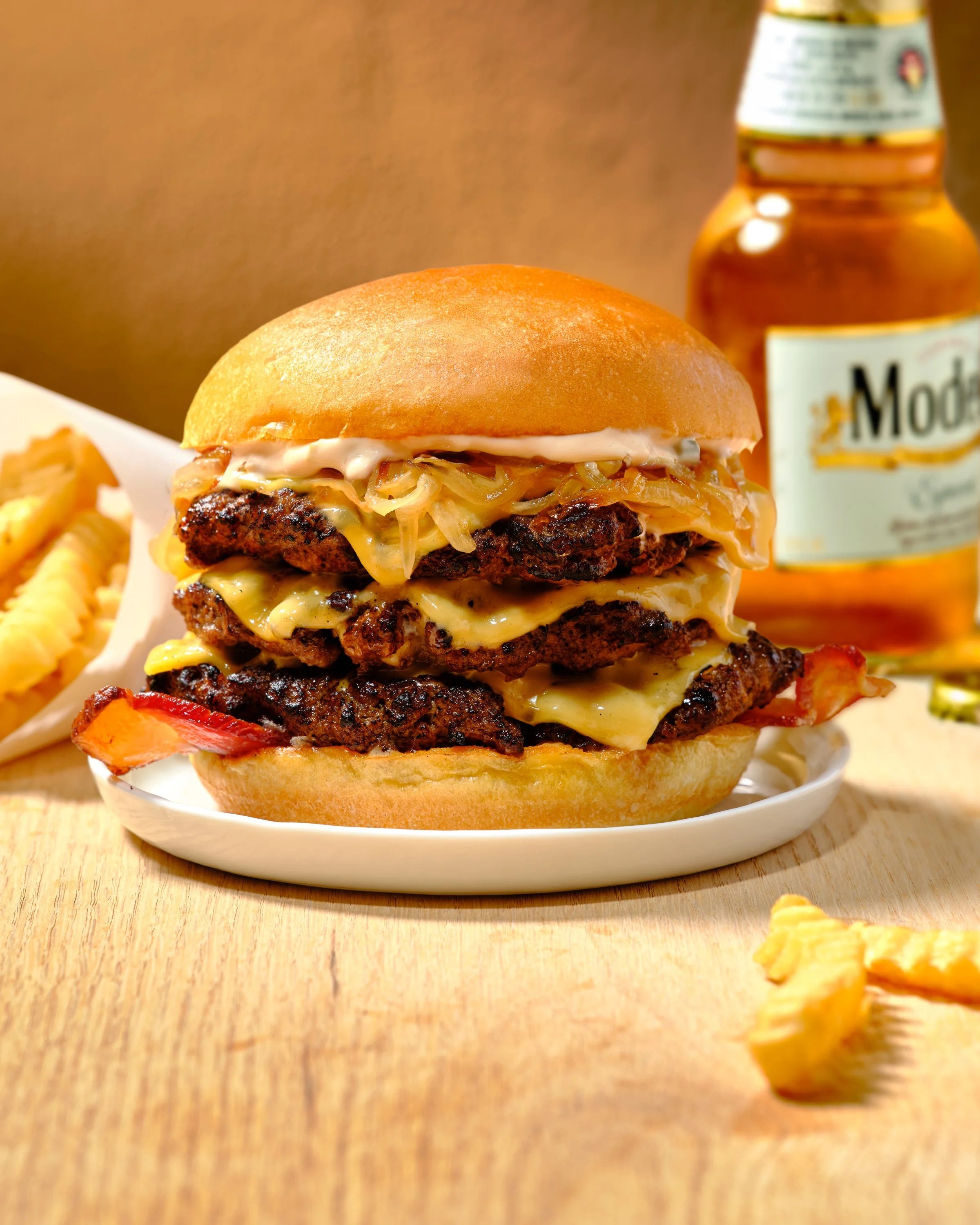 A large triple patty cheeseburger with melted cheese, caramelized onions, bacon, and sauce in a bun, served on a white plate with fries and a bottle of hot sauce on a wooden table. Chicago Milwaukee Midwest Illinois Food Food beverage Food Drink Savo