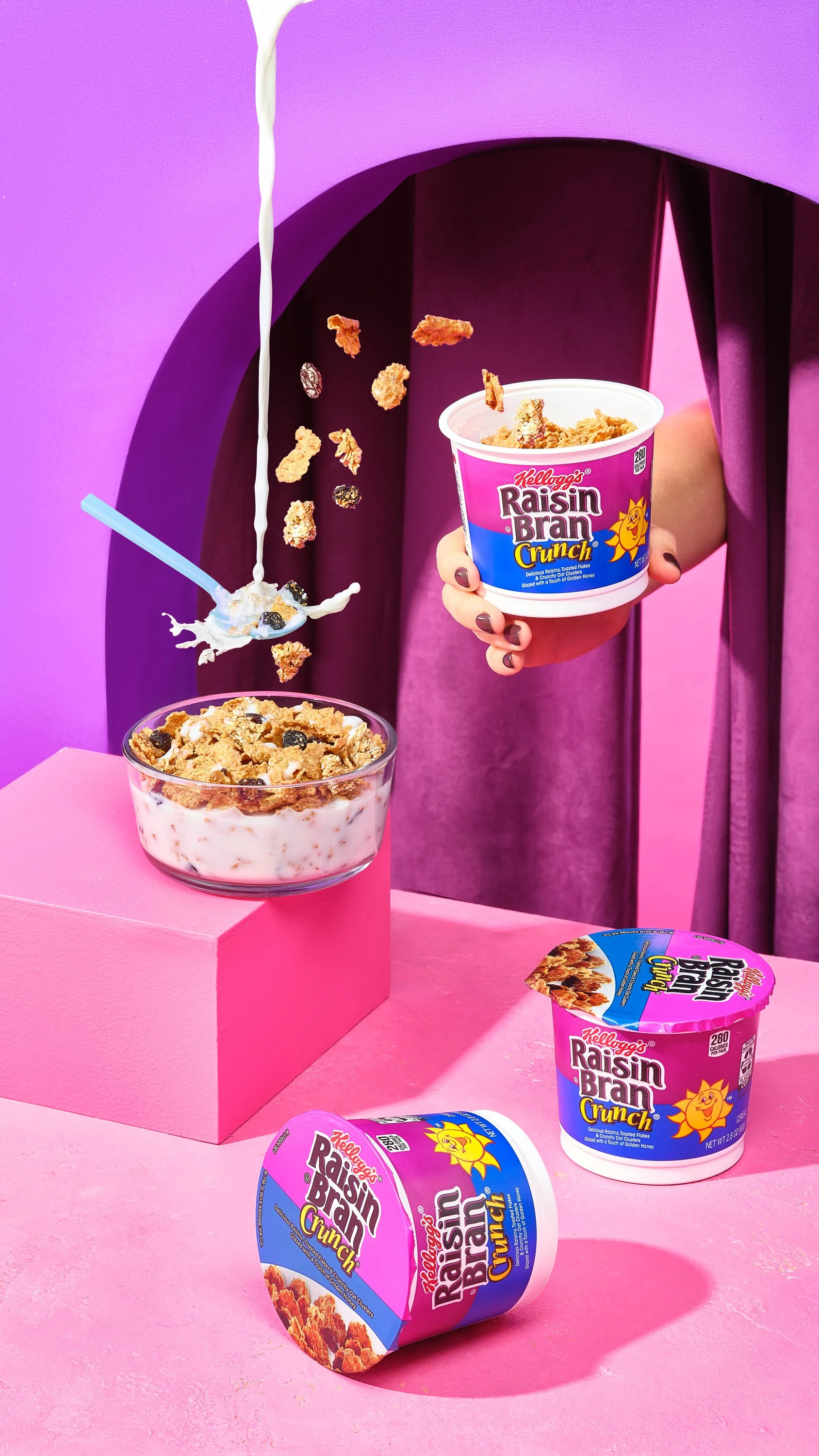 A person holding a cup of Kellogg's Raisin Bran Crunch cereal, with cereal and milk being poured into a glass bowl, and other cereal containers on a pink surface with a purple background. Chicago Milwaukee Midwest Illinois Food Food beverage Food Dri