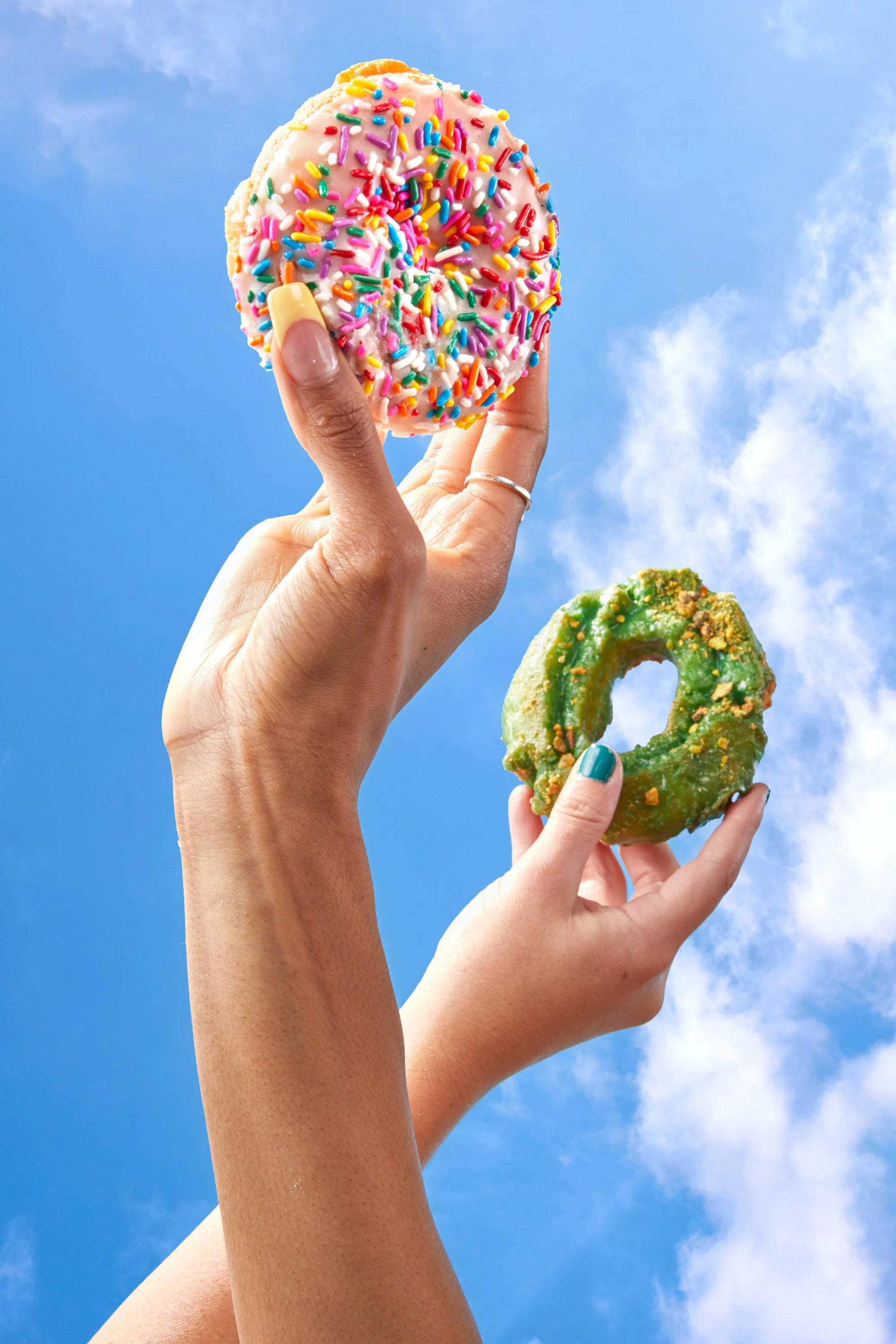 Two hands holding colorful donuts against a blue sky with clouds. Chicago Milwaukee Midwest Illinois Food Food beverage Food Drink Savory Photographers photographer photography videography videographer CPG Bennett Fuhrman Ben Tiny Scheme