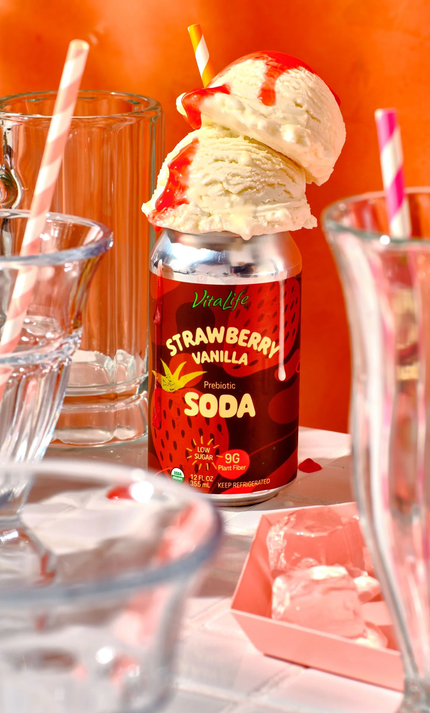 A can of strawberry vanilla prebiotic soda topped with two scoops of vanilla ice cream drizzled with strawberry syrup, surrounded by glasses with striped straws and a pink box with marshmallows, set against an orange background.