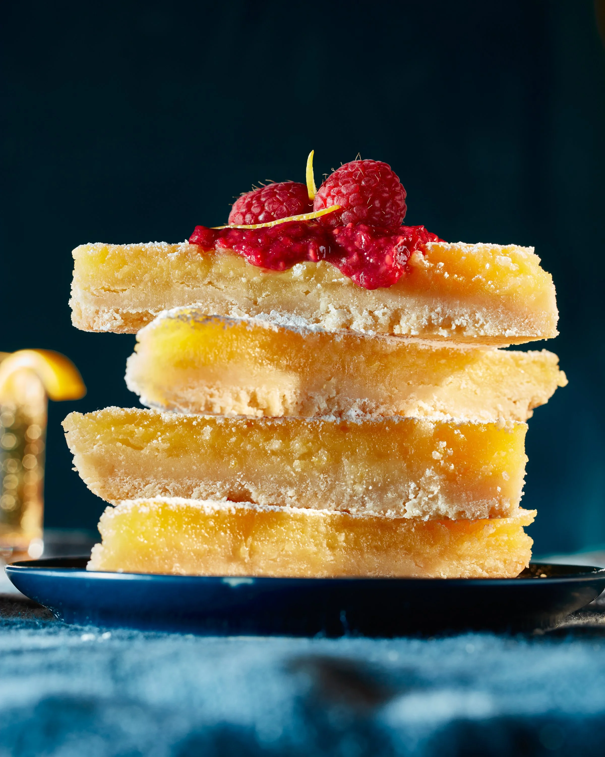 Stack of four lemon bars topped with raspberries and lemon zest on a dark background. Chicago Milwaukee Midwest Illinois Food Food beverage Food Drink Savory Photographers photographer photography videography videographer CPG Bennett Fuhrman Ben Tiny