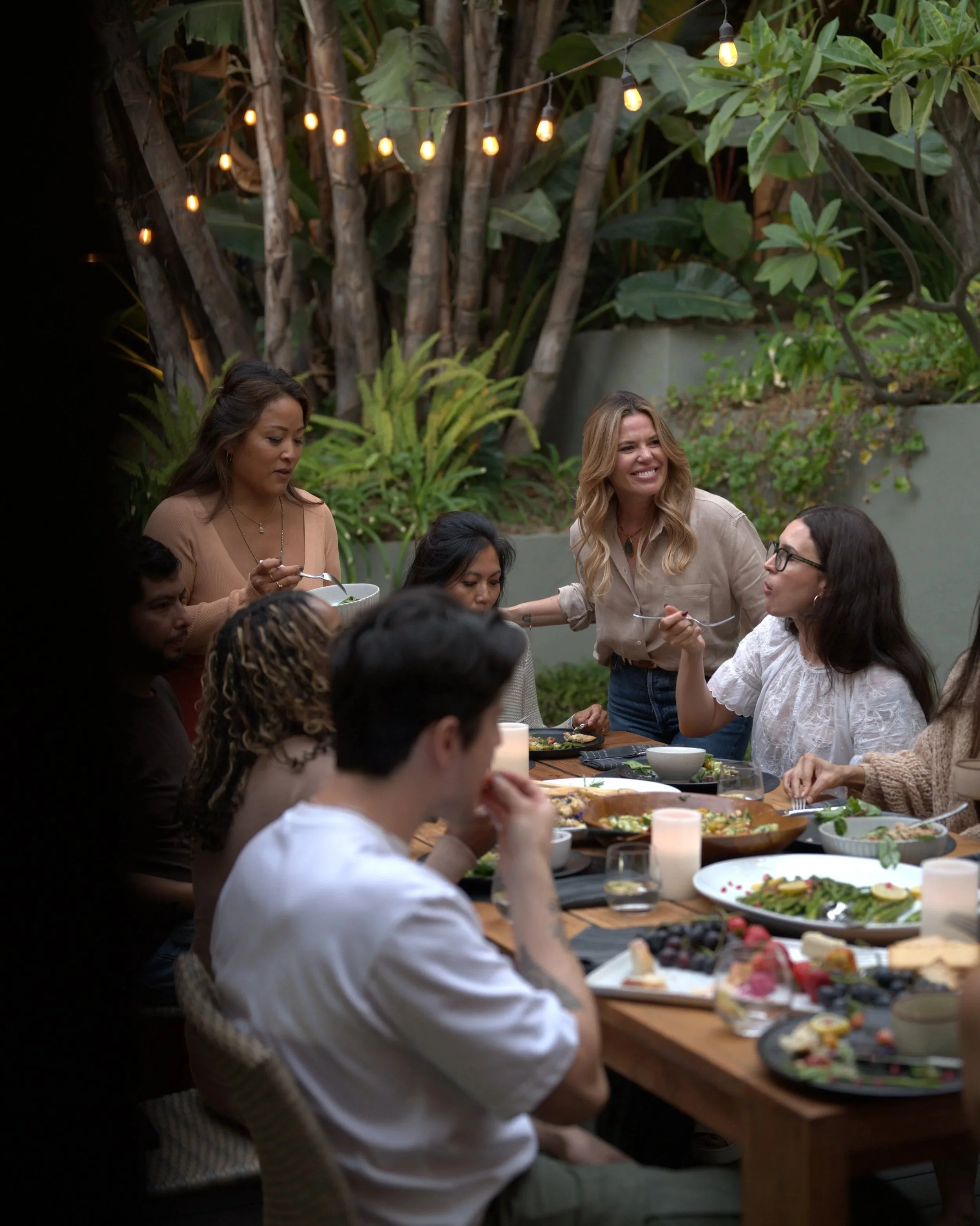 People gathered around a dinner table outdoors, eating and chatting, with string lights and lush greenery in the background.