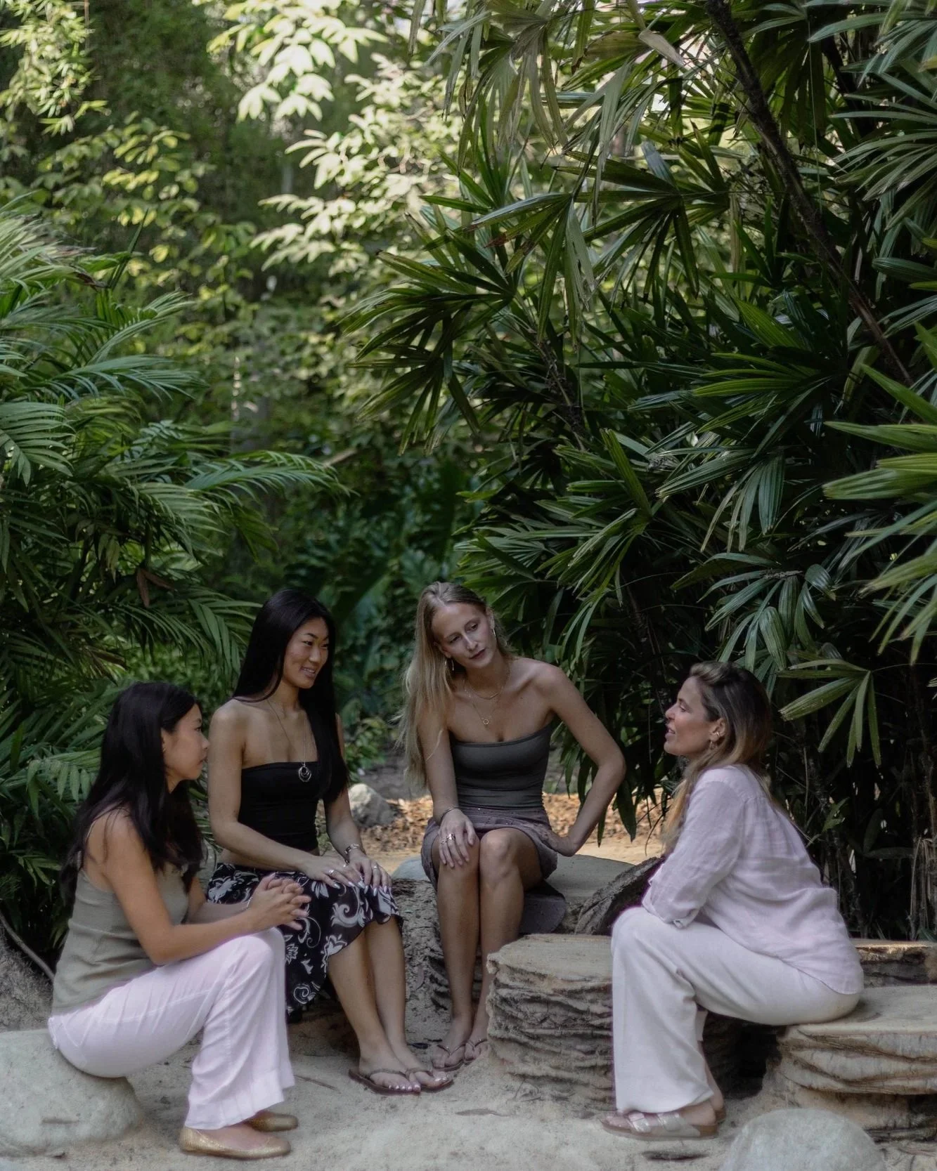 Four women sitting outdoors on rocks among lush green tropical plants, engaged in conversation.