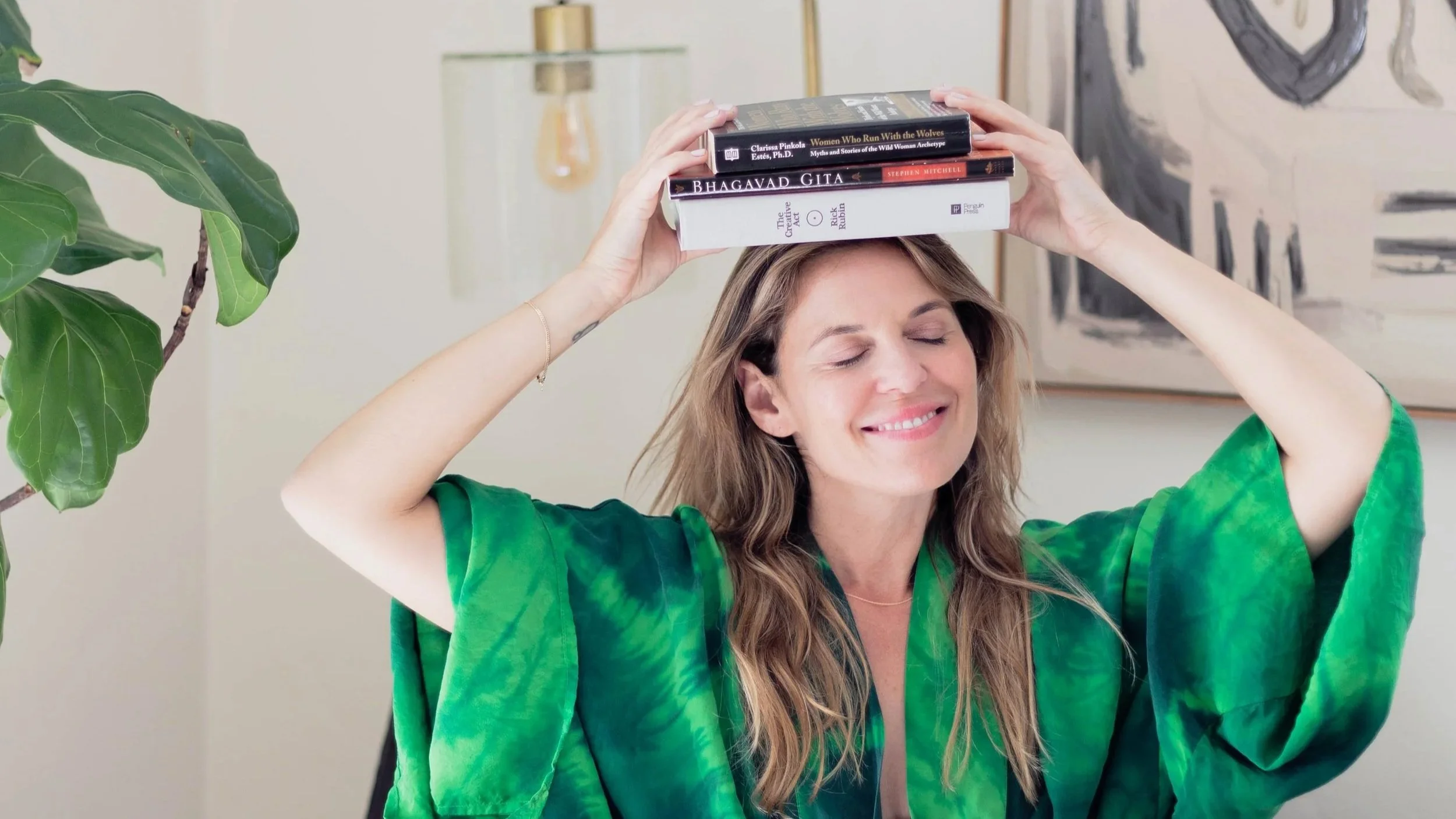 Dr. Courtnay Meletta smiling with eyes closed, balancing a stack of books on her head, seated indoors near a green plant and abstract art in the background.