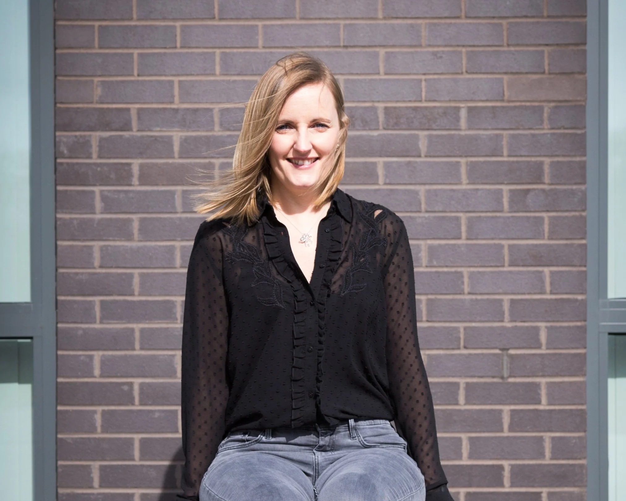 Person with blonde hair wearing a black shirt and gray jeans, sitting against a brick wall background.
