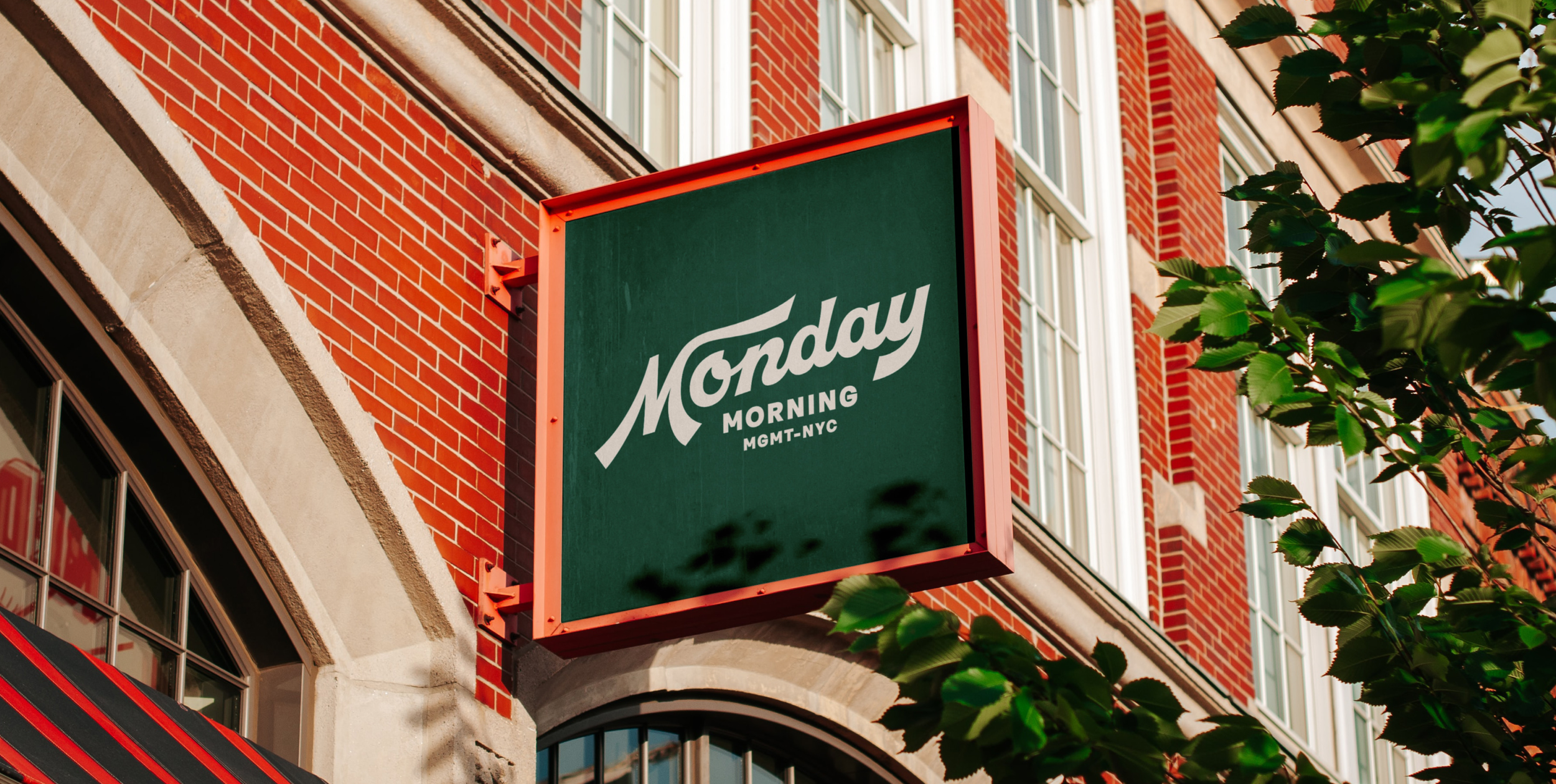 Green hanging sign reading 'Monday Morning MGMT-NYC' in front of a red brick building with large windows, partly obscured by green leaves.