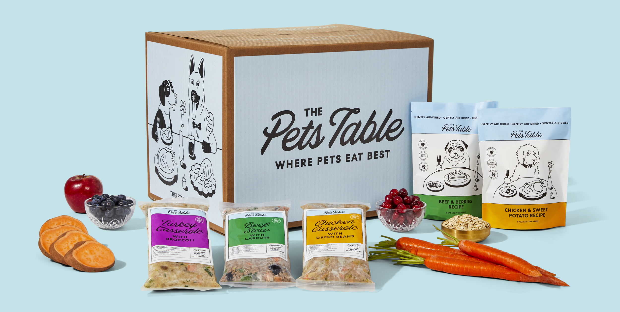 Assorted pet food products and ingredients arranged around a large blue cardboard box labeled "The Pets Table" with the tagline "Where Pets Eat Best".