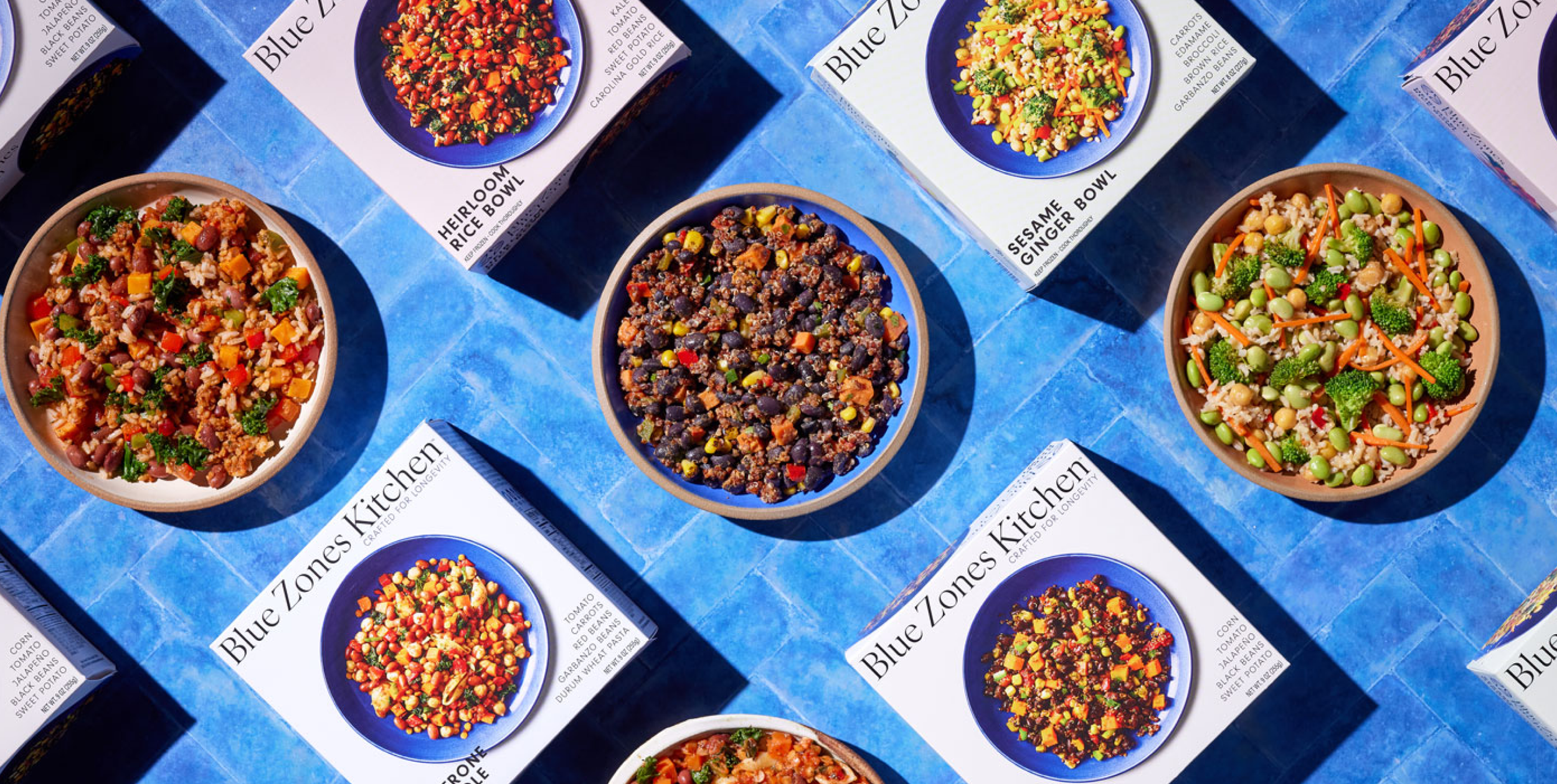Multiple boxes of Blue Zones meal kits with bowls of colorful vegetarian salads on a blue tiled surface.