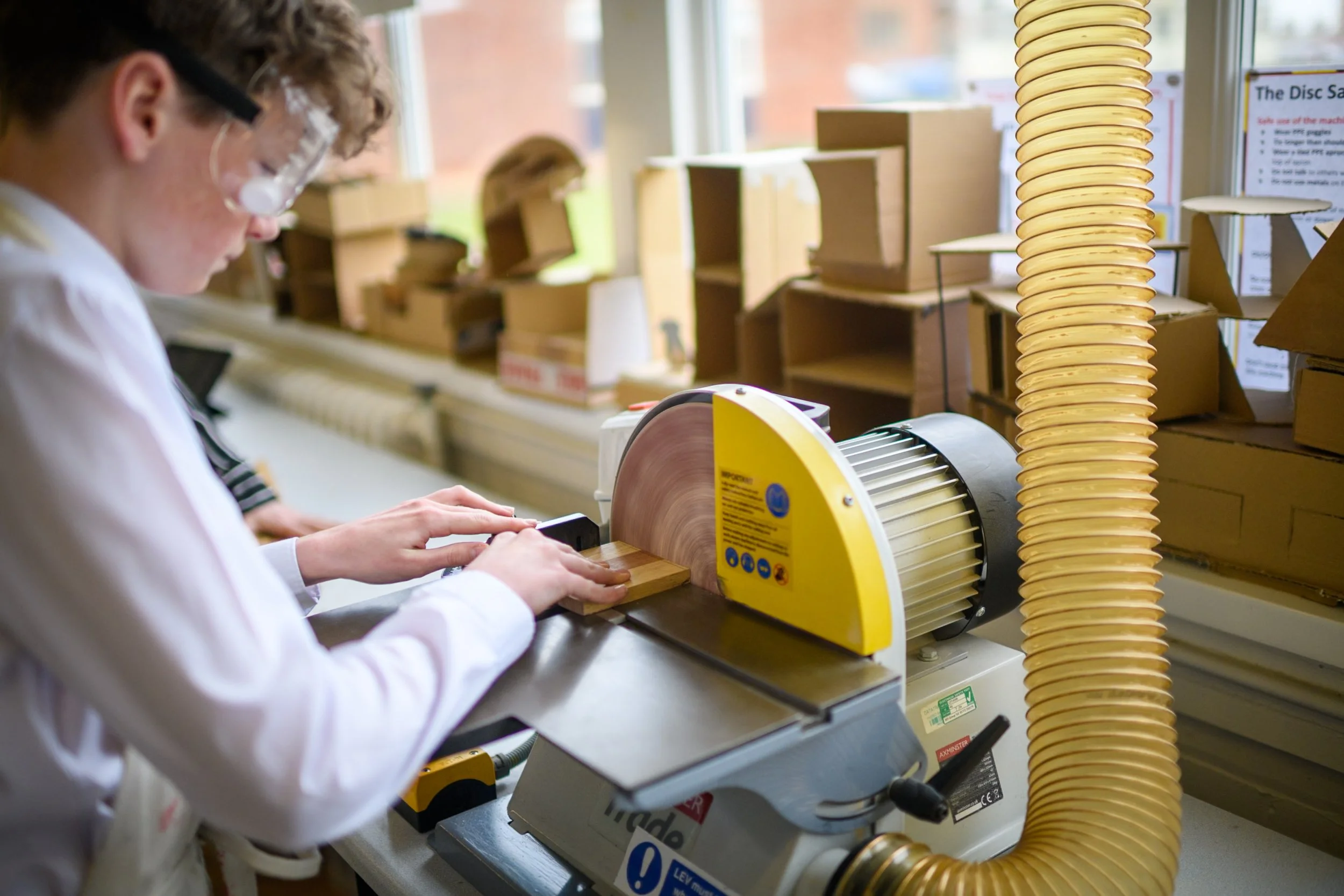 A Rossall School student working with a wood disc cutting machine in the DT workshop, wearing safety goggles, with wooden shelves and boxes in the background.