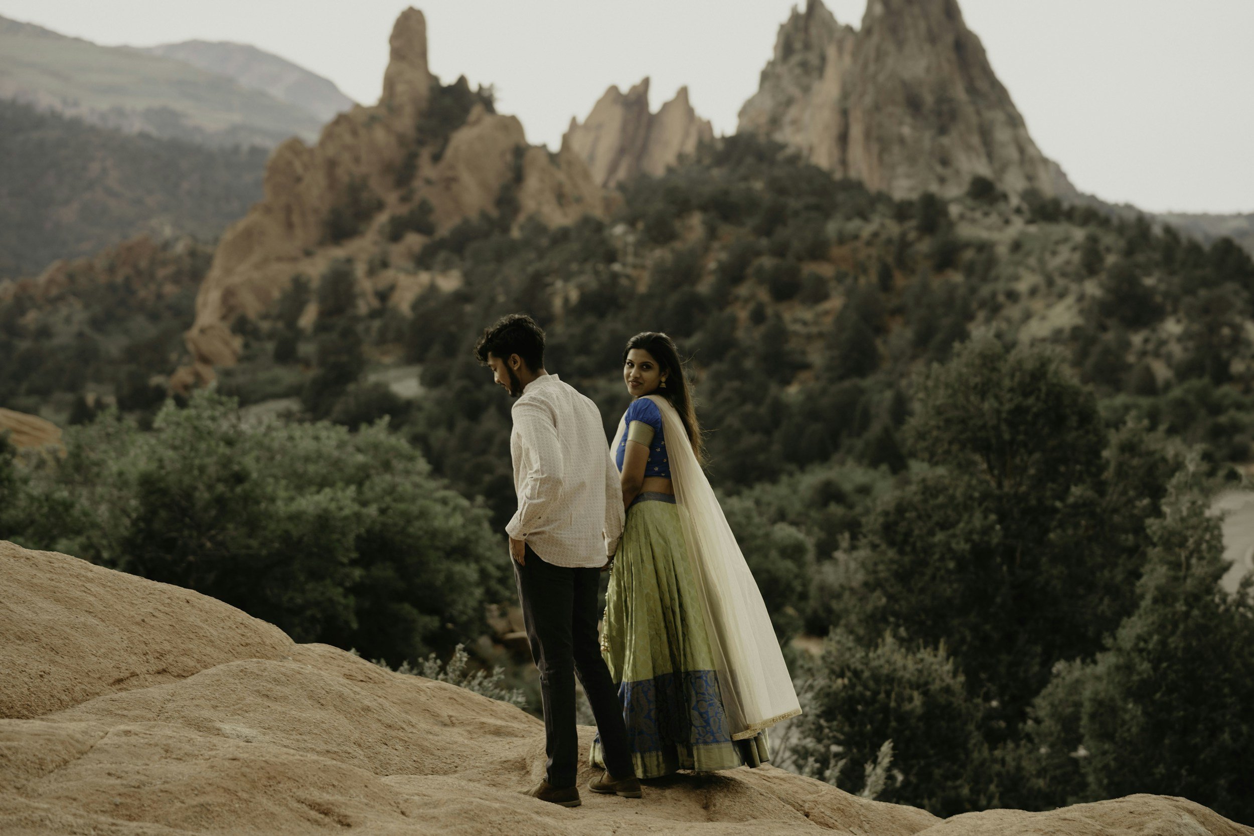 A man and woman standing on rocky terrain with mountainous and forested landscape in the background. The woman wears a traditional outfit with a blue top and green skirt, while the man wears a light-colored shirt and dark pants.