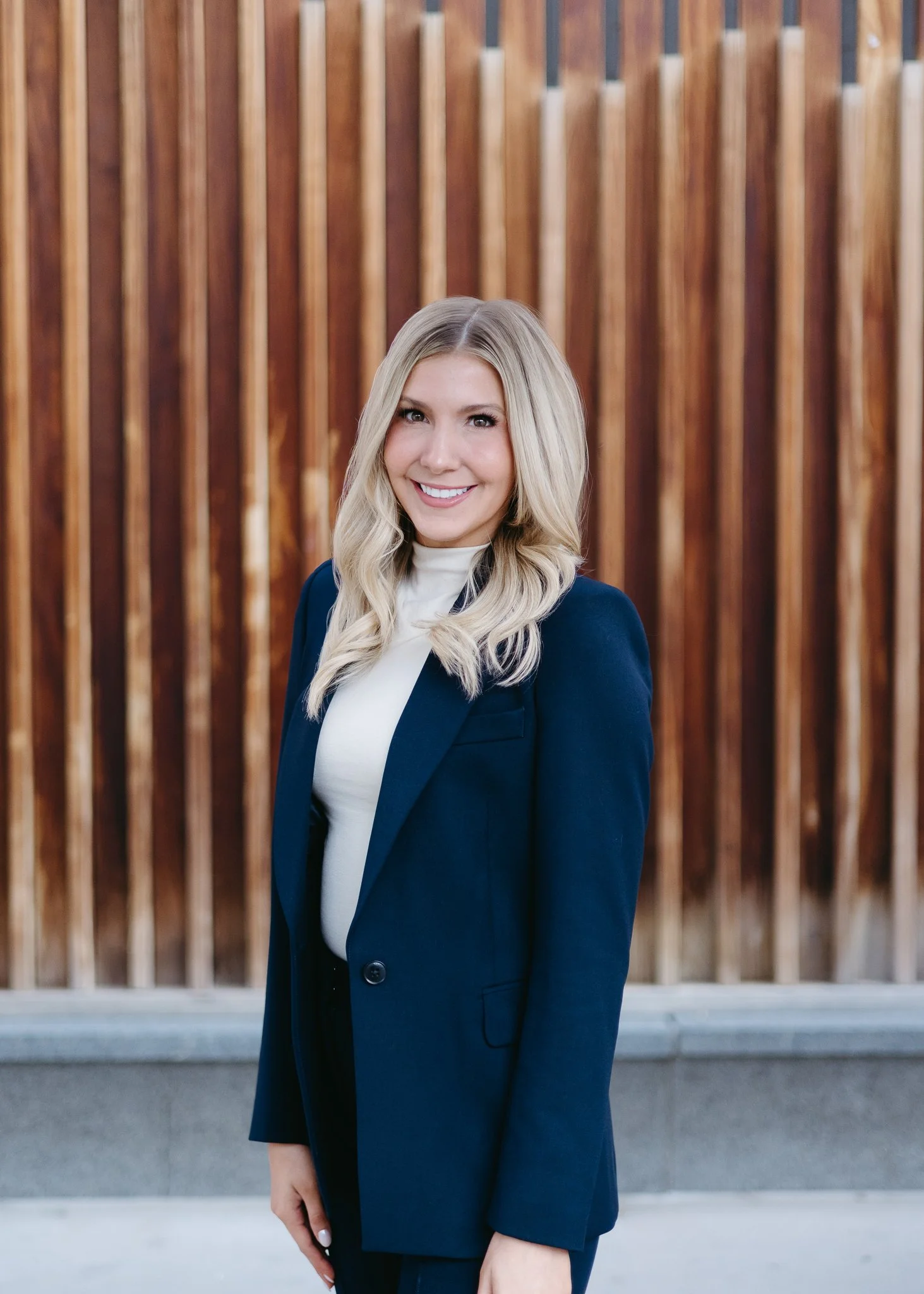 A woman with blonde hair wearing a navy blazer and white top standing in front of a wooden fence, smiling at the camera.