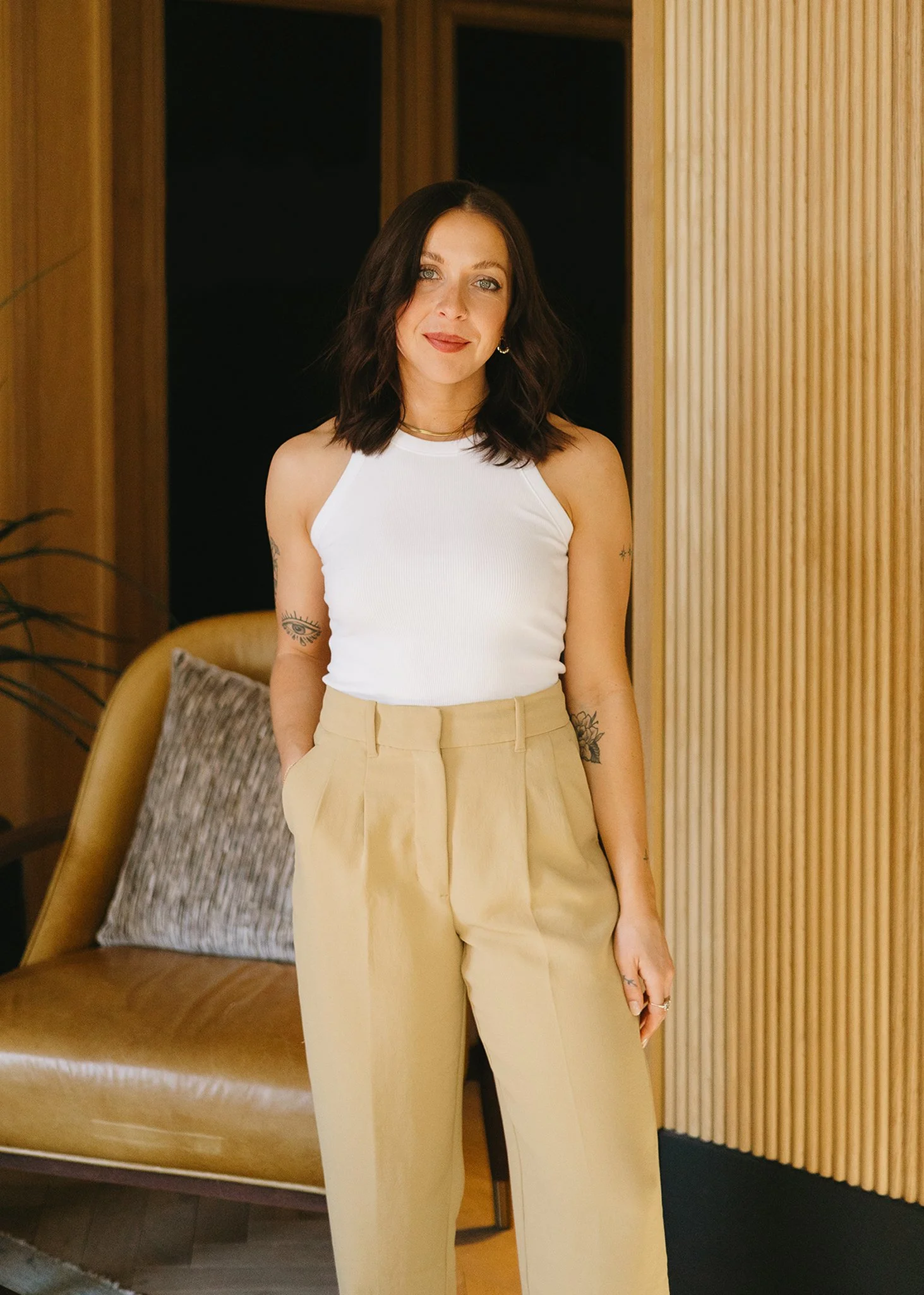 A woman with shoulder-length dark hair and tattoos on both arms, wearing a white sleeveless top and beige high-waisted trousers, standing indoors in front of a wooden wall, with a leather armchair and pillow in the background.