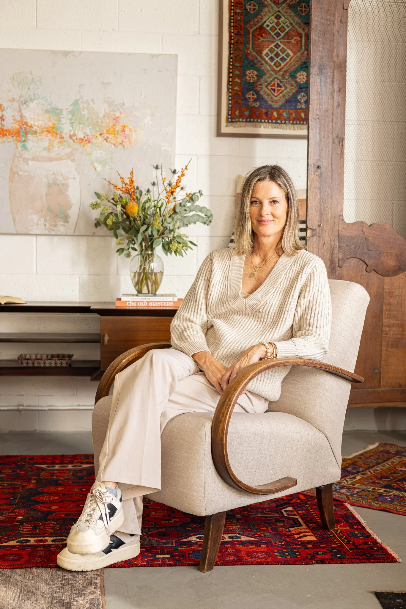 A woman sitting on a beige armchair with wooden armrests in an art-filled room, with a large bouquet of flowers on the table behind her and a colorful rug on the floor.
