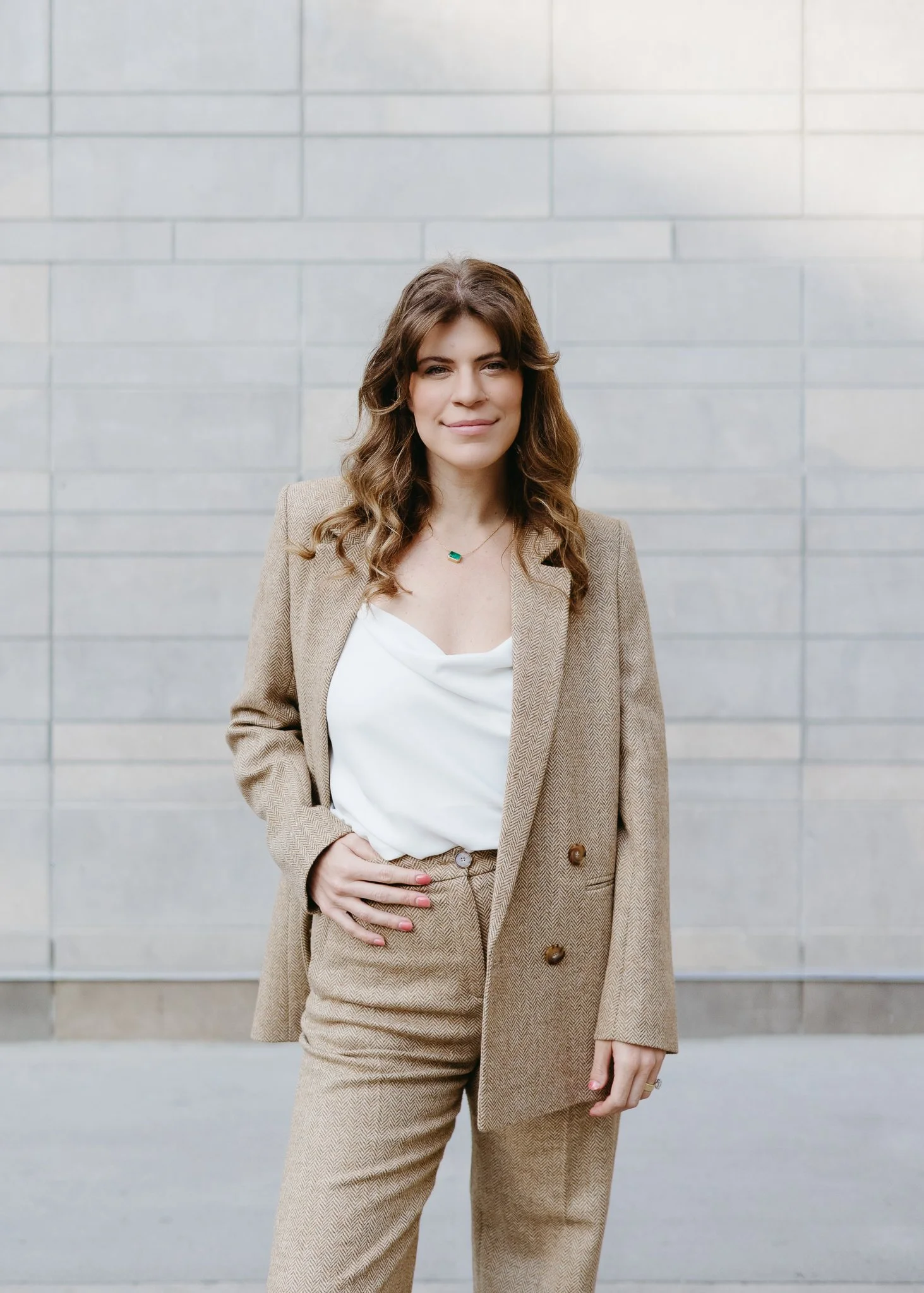 A woman standing outside against a gray brick wall, wearing a beige suit and white top, with wavy brown hair and a slight smile.